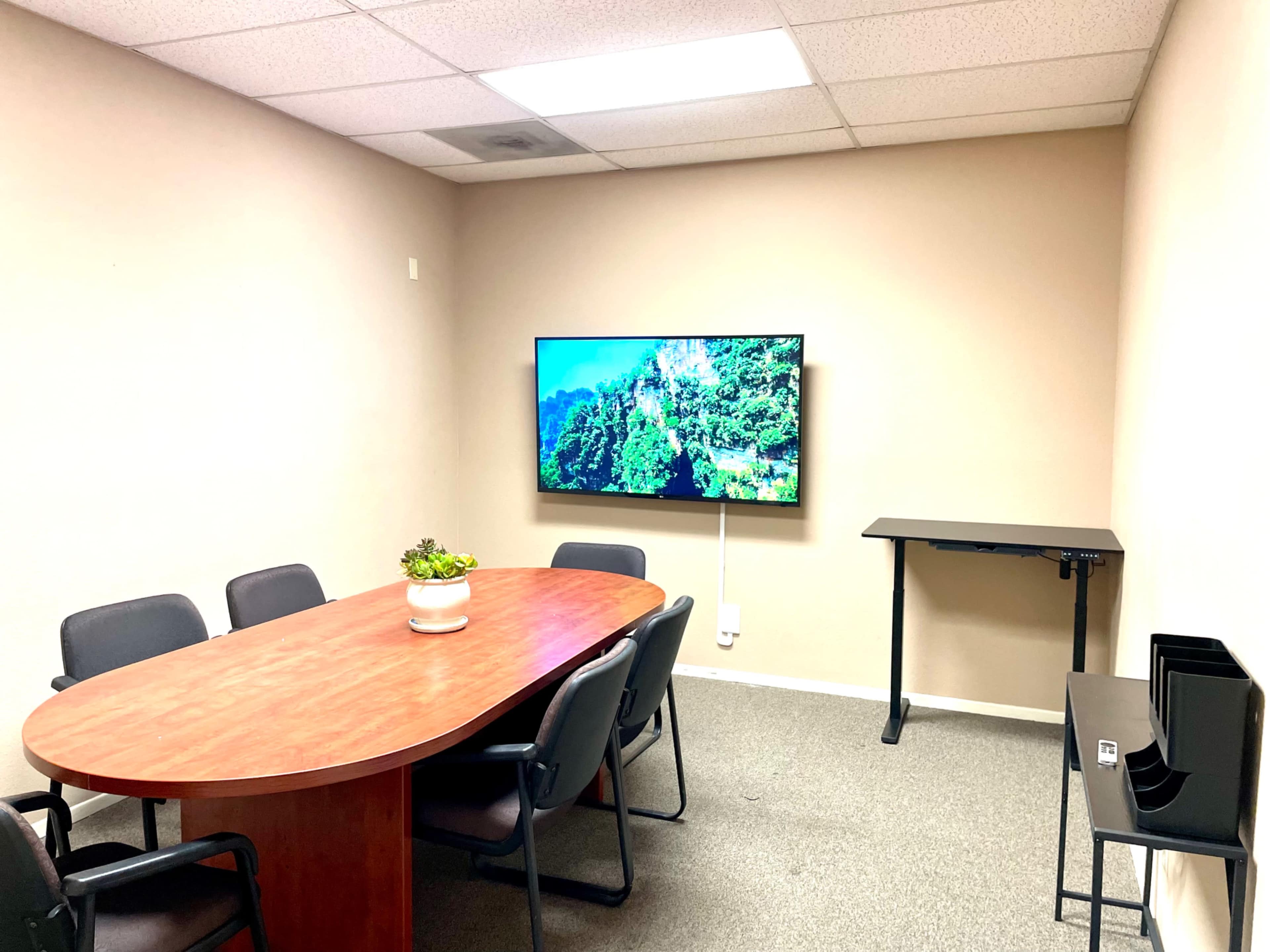 A small conference room with a wooden oval table, six black chairs, a mounted television displaying a nature scene, and a standing desk against one wall.