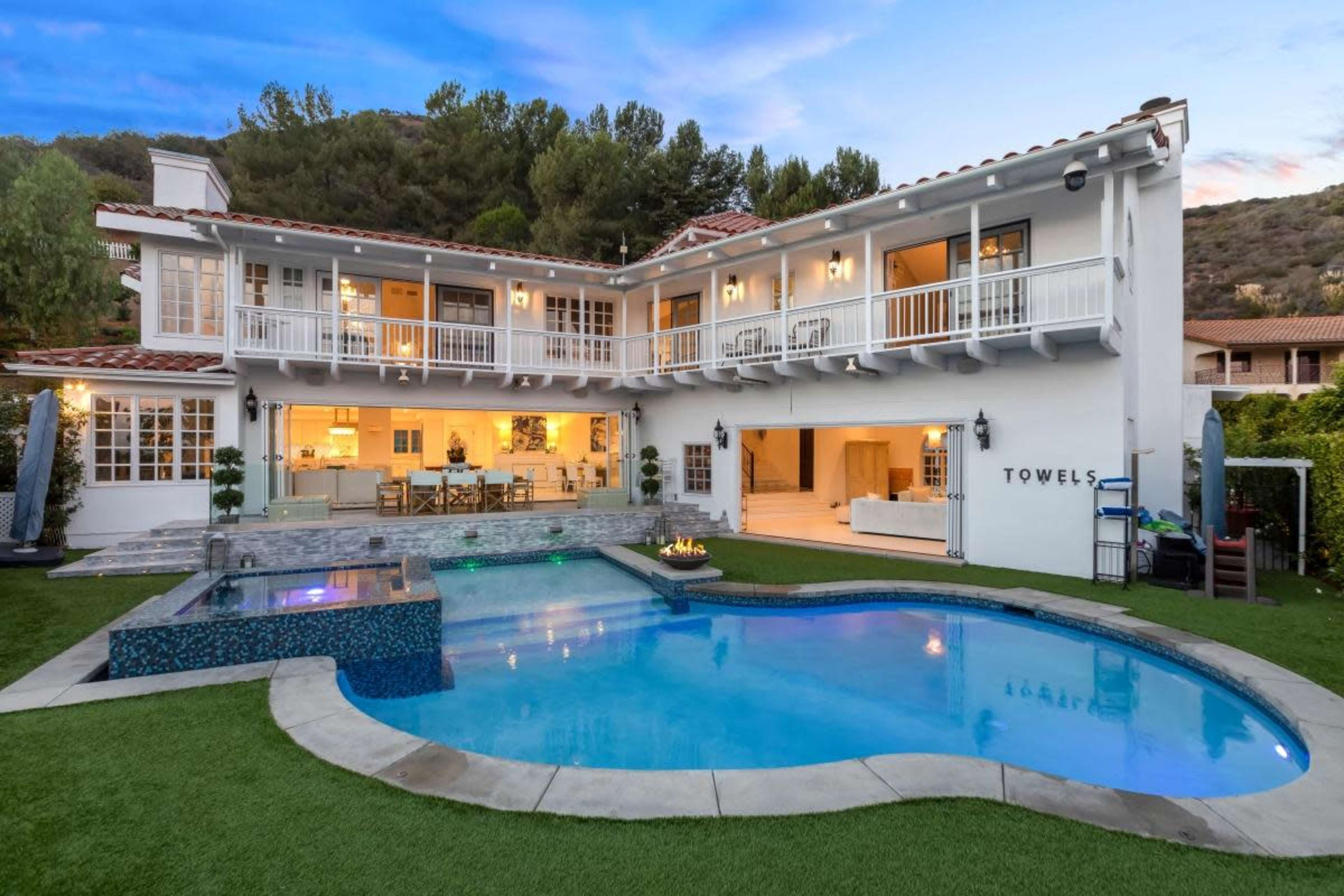 The image shows a two-story house with a swimming pool and outdoor seating area, set against a hillside backdrop.