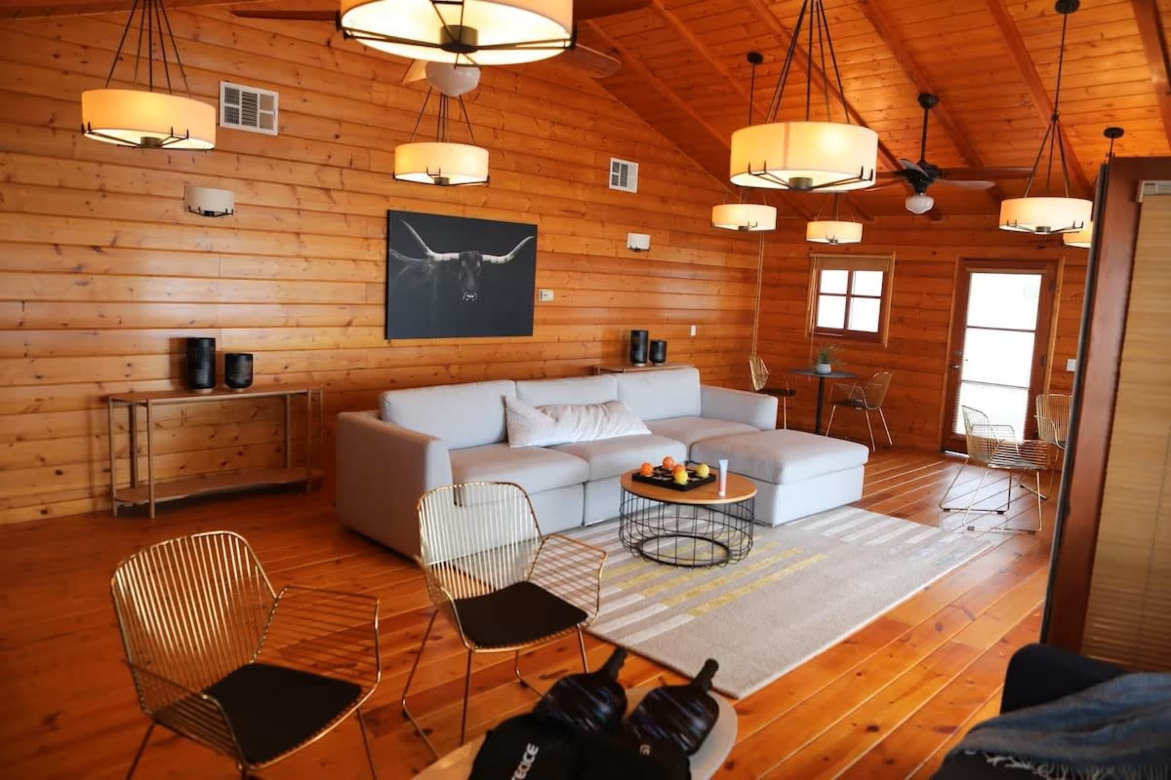 A cozy wooden cabin living room with a large gray sectional sofa, a coffee table with snacks, and metal chairs arranged around a light-colored rug.