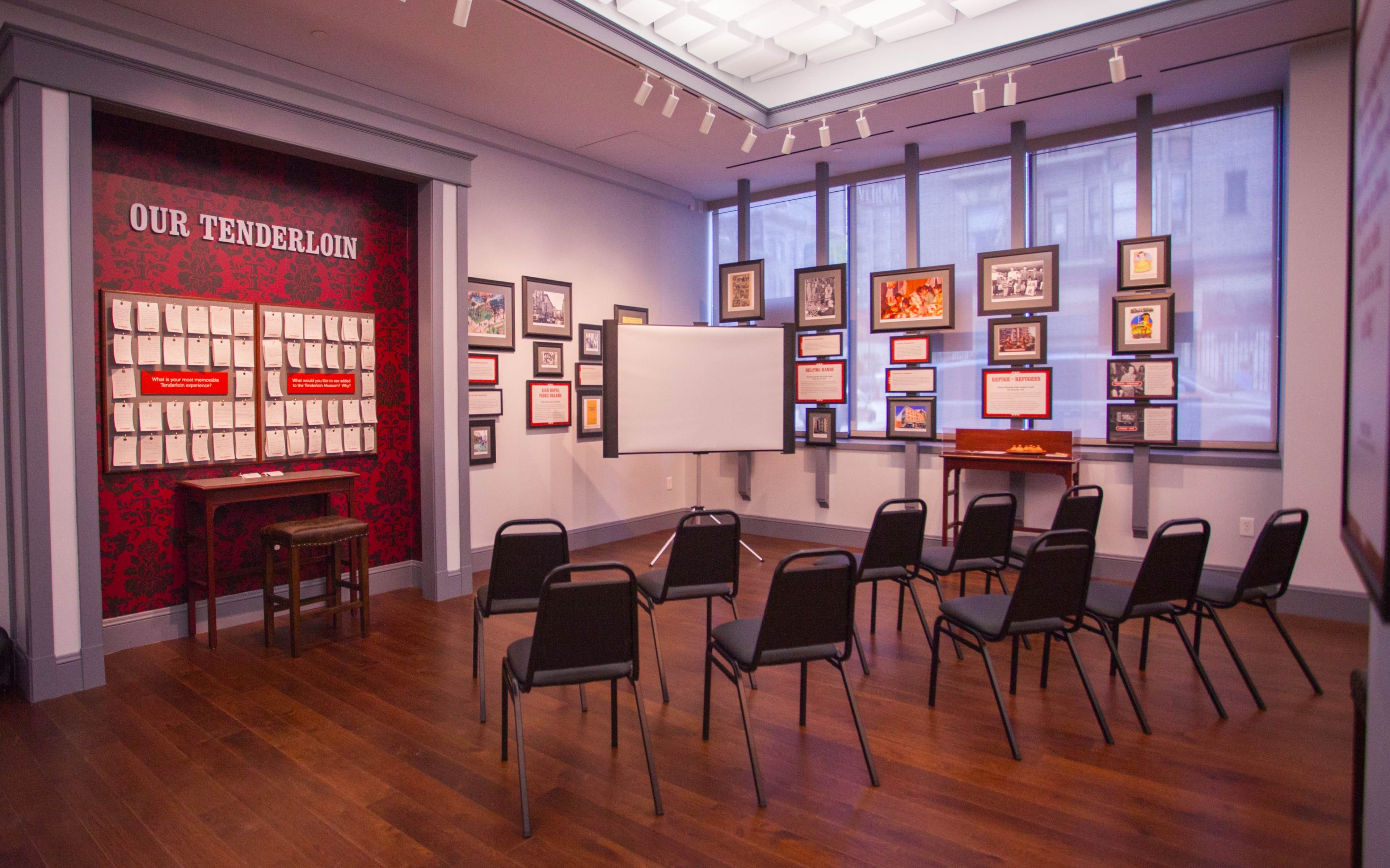 A room set up for a presentation, featuring a blank screen, several framed pictures on the walls, and chairs arranged in a semicircle facing the screen.