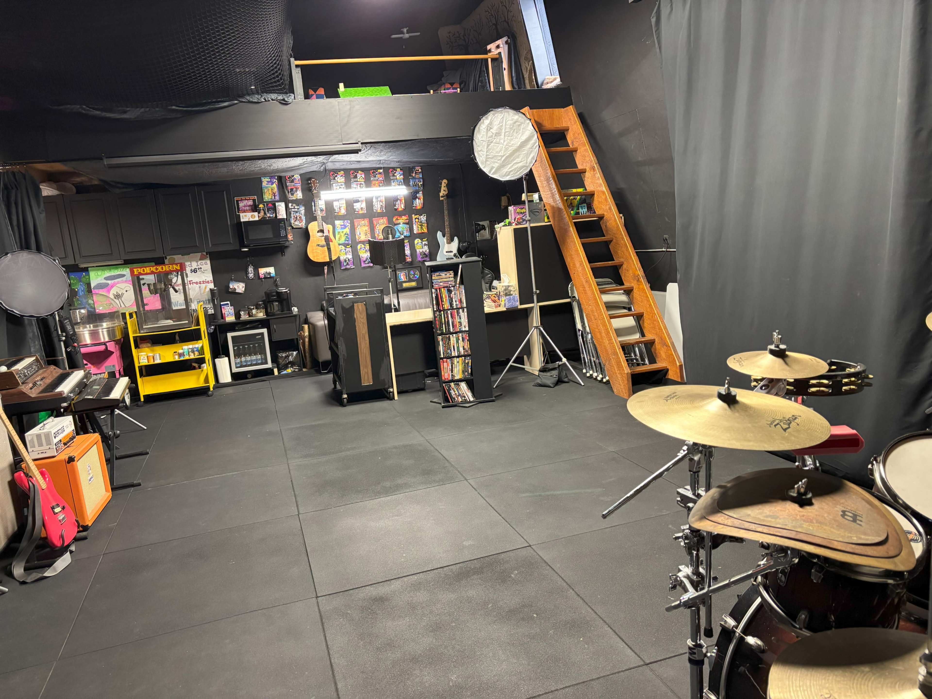 The image shows a music practice space with a drum set in the foreground, shelves of instruments and equipment along the walls, and a staircase leading to a second level.