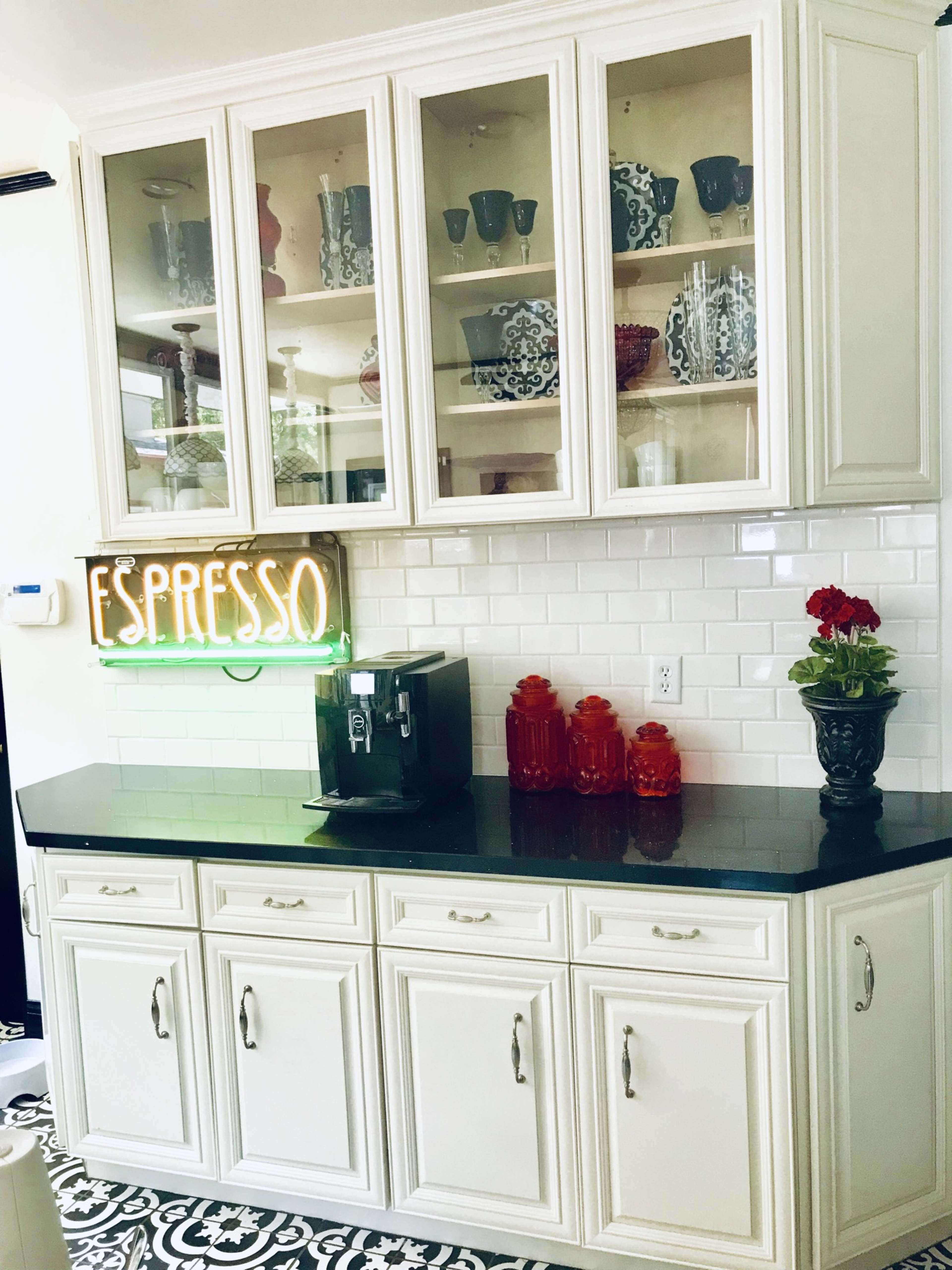 A kitchen countertop features a coffee machine, decorative red glassware, and a neon "ESPRESSO" sign against white cabinets and a black countertop.