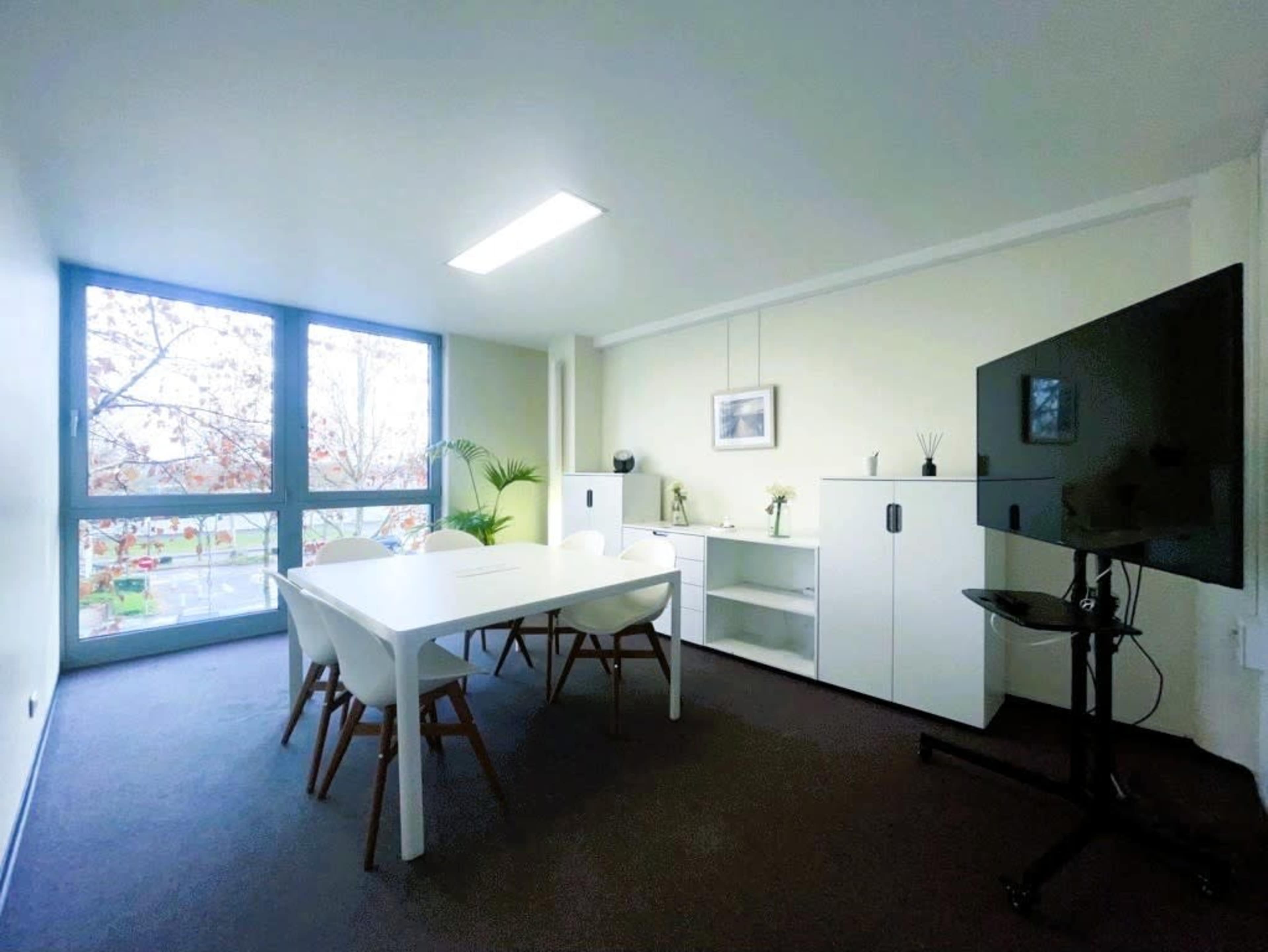 The image shows a modern meeting room featuring a white table with chairs, a storage cabinet, and a television mounted on a stand, with large windows letting in natural light.