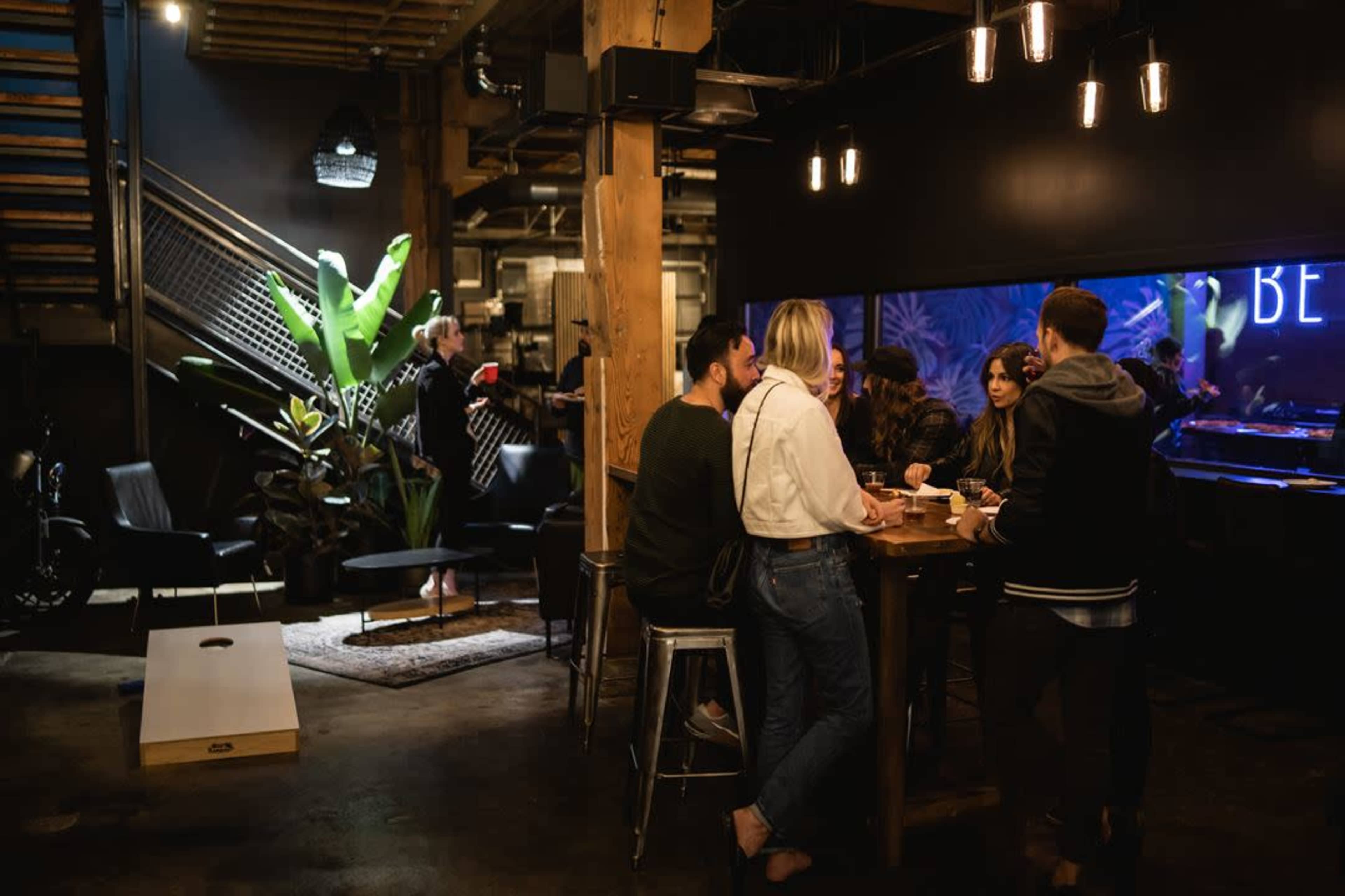 A group of people converse around a wooden table in a modern cafe with industrial decor and plants.