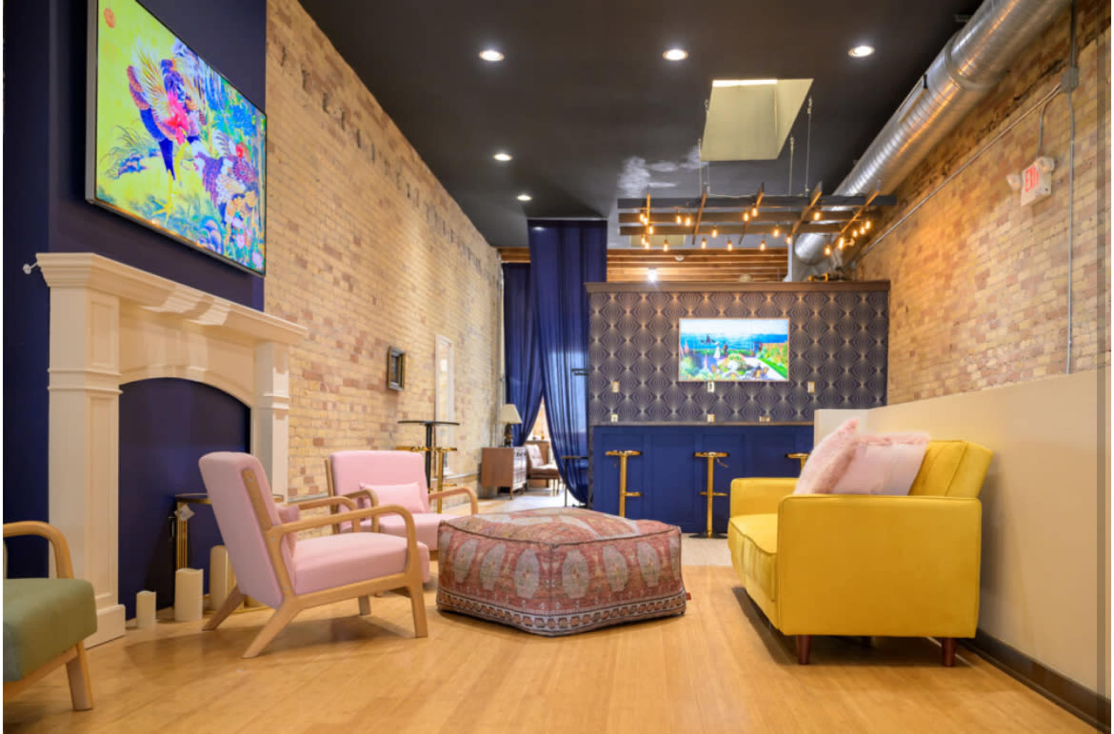 The image shows a modern interior living space featuring a yellow sofa, pink armchairs, a patterned ottoman, and wall-mounted televisions with a blend of exposed brick and stylish decor.