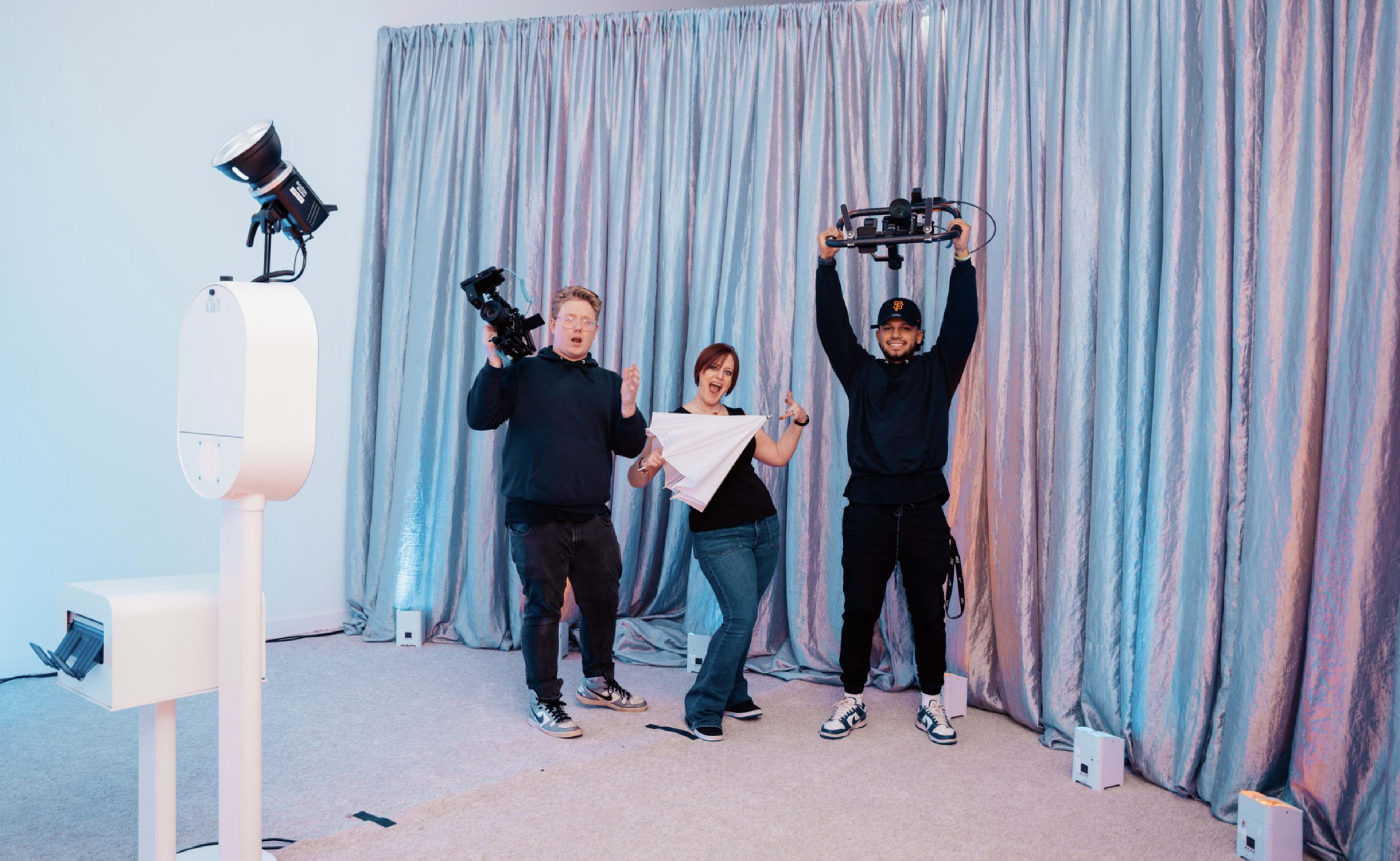Three individuals are smiling and posing in a studio setting with a backdrop of metallic curtains, one holding a camera, another holding a drone, and the third standing between them.