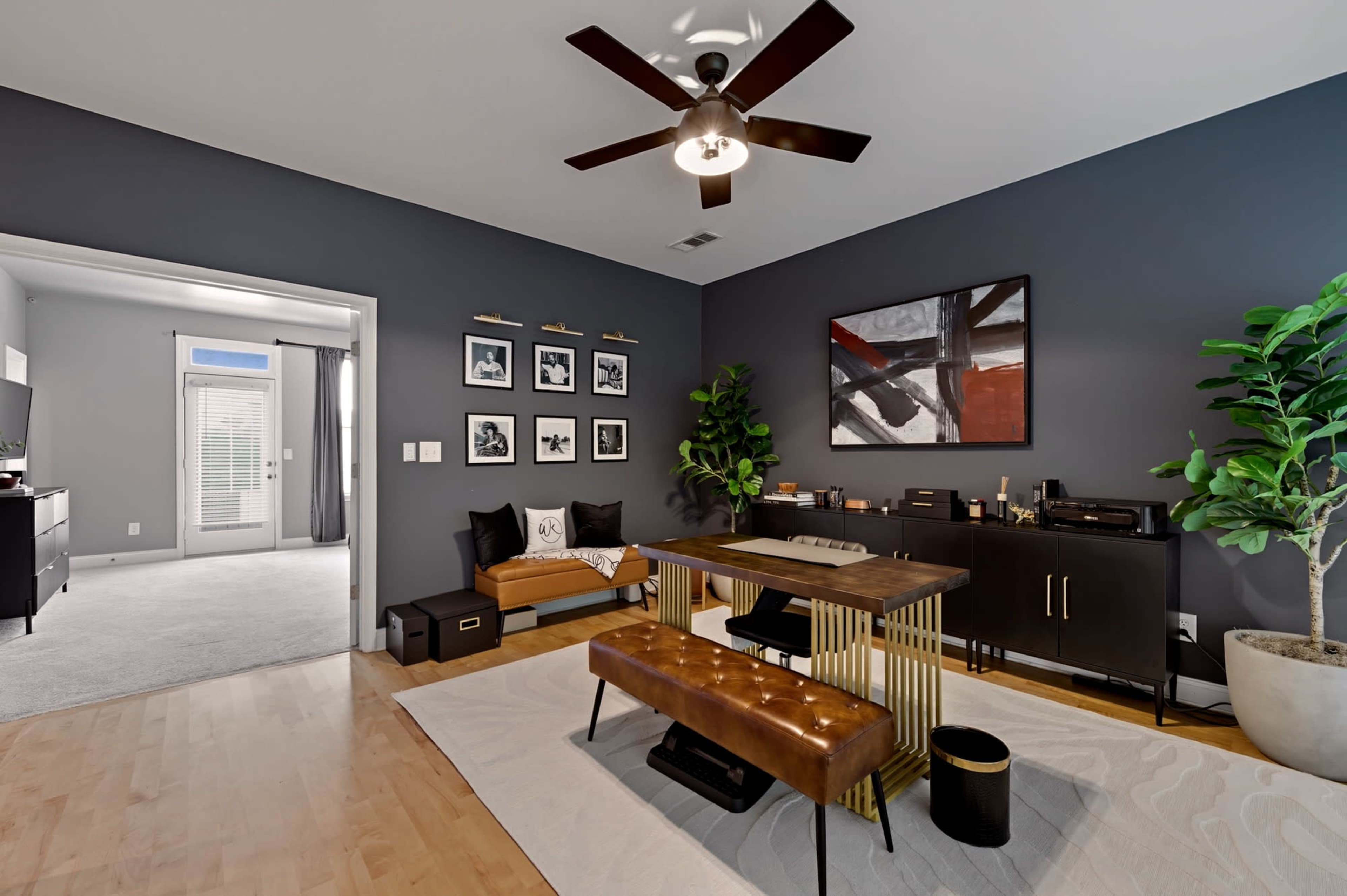 The image shows a modern office space with a dark wall, a wooden desk, a bench, and various framed artworks.