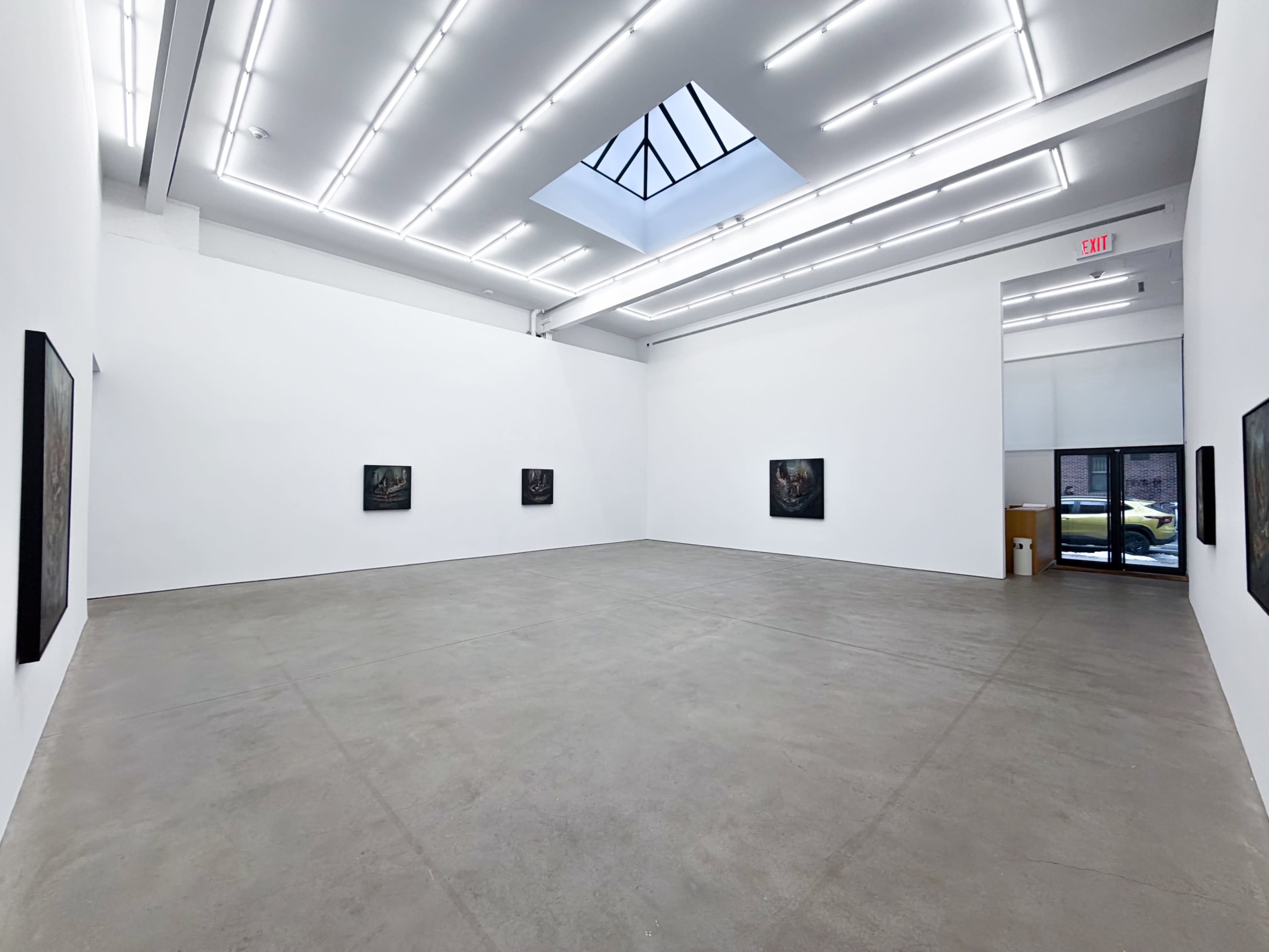 The image shows a spacious, modern art gallery with four dark-framed paintings displayed on the white walls, illuminated by overhead lights and a skylight.