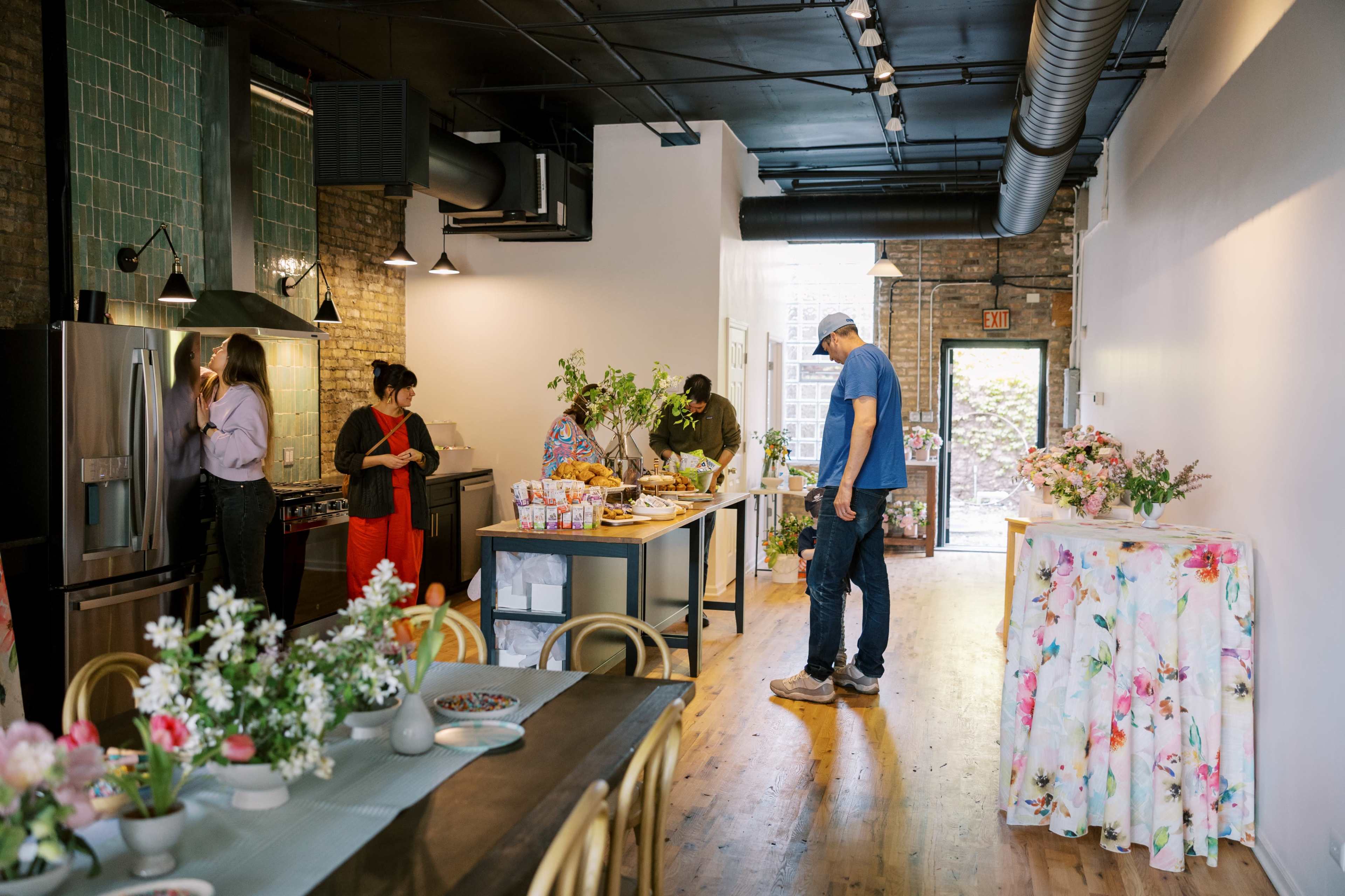 A modern kitchen and dining area is set up for an event, featuring a buffet table with food, a floral arrangement, and guests interacting.