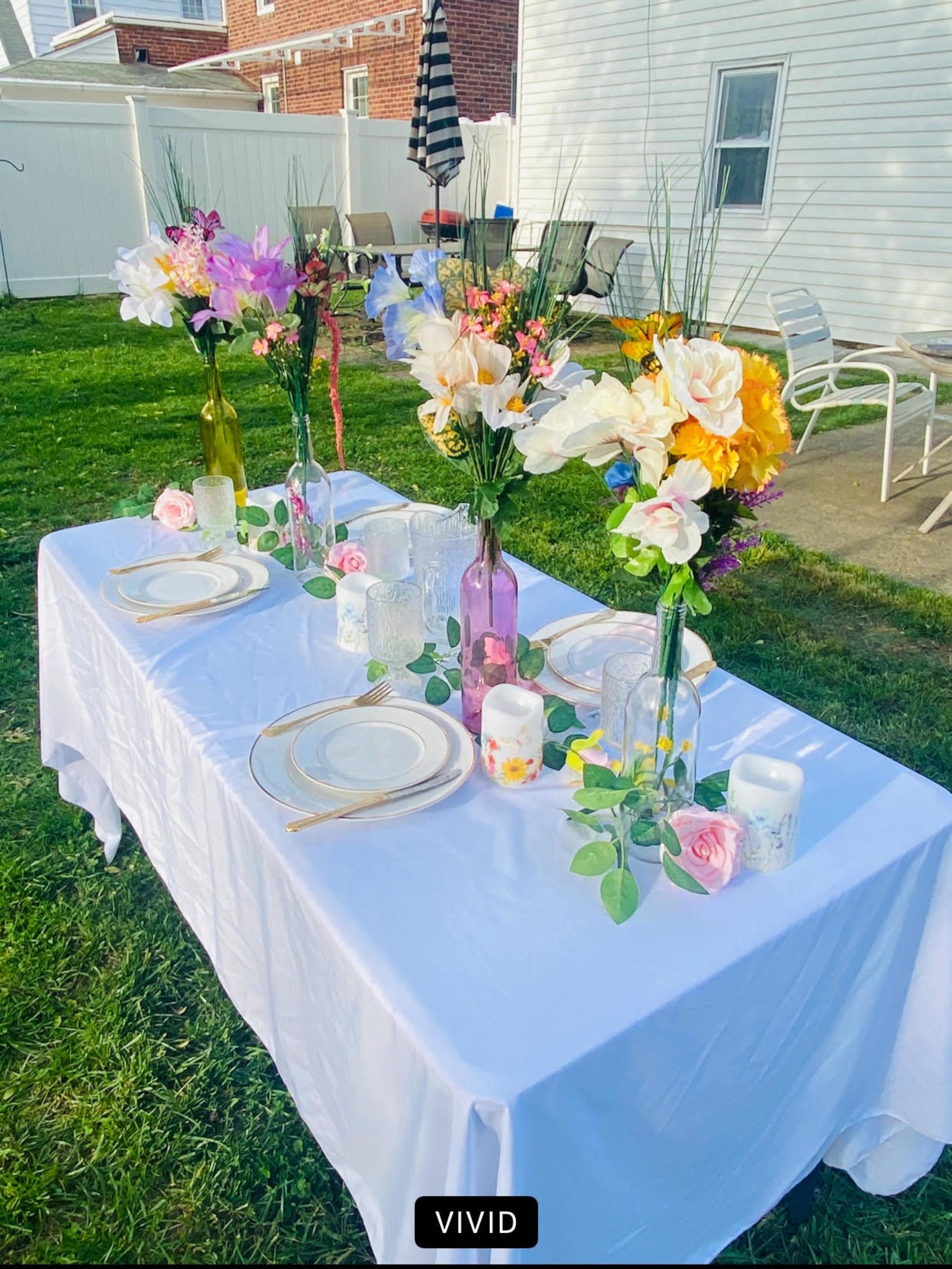 A table is set outdoors with a white tablecloth, colorful flower arrangements in vases, plates, and decorative candles.