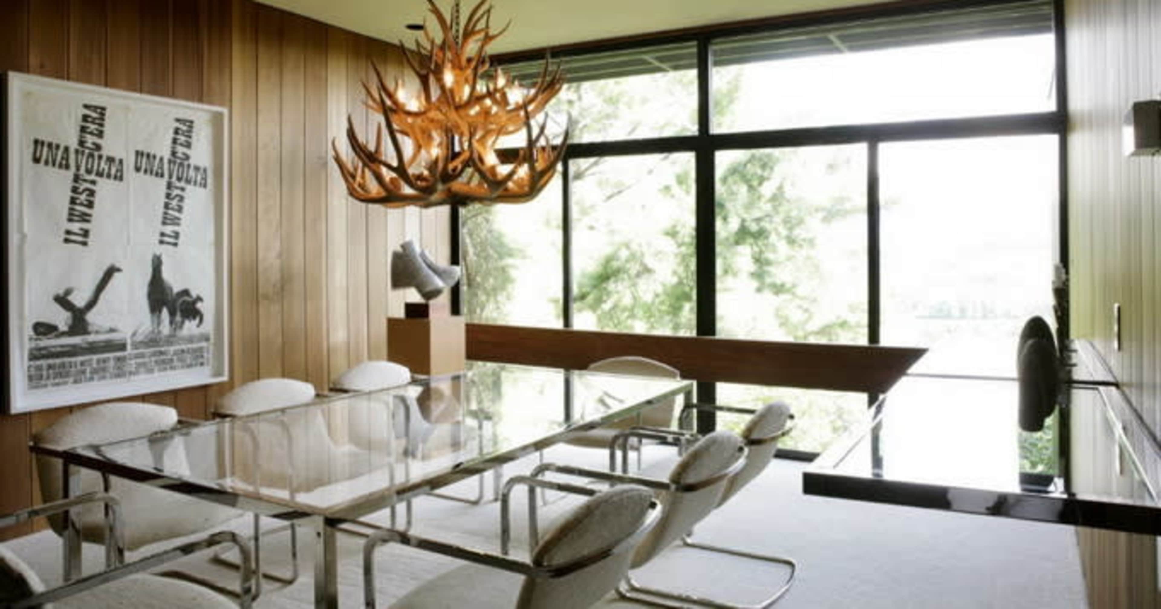 A modern dining room features a glass table surrounded by metal chairs, large windows letting in natural light, and an artistic antler chandelier hanging from the ceiling.