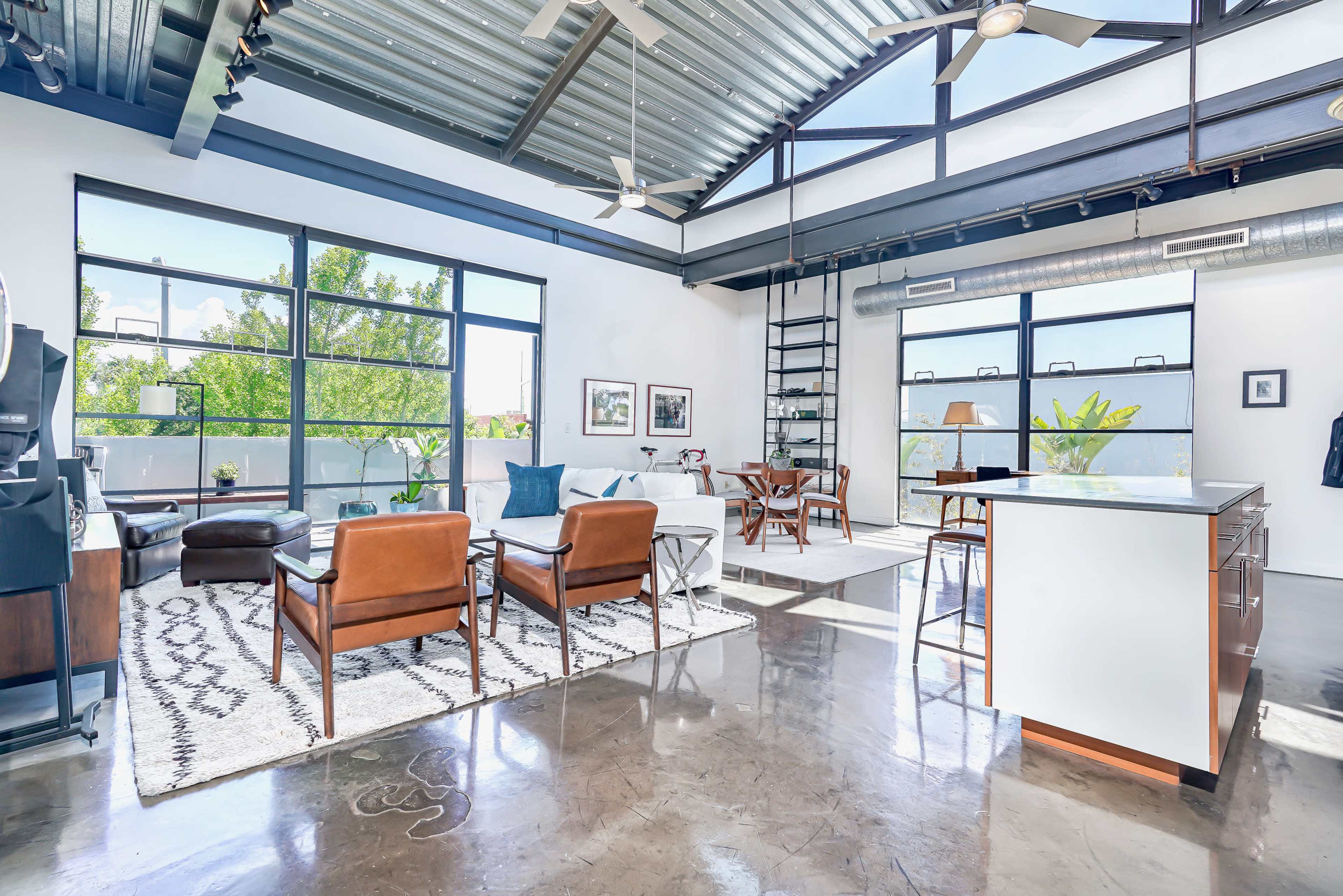 The image shows a spacious, modern living area with large windows, polished concrete floors, and a mix of comfortable seating and dining options.