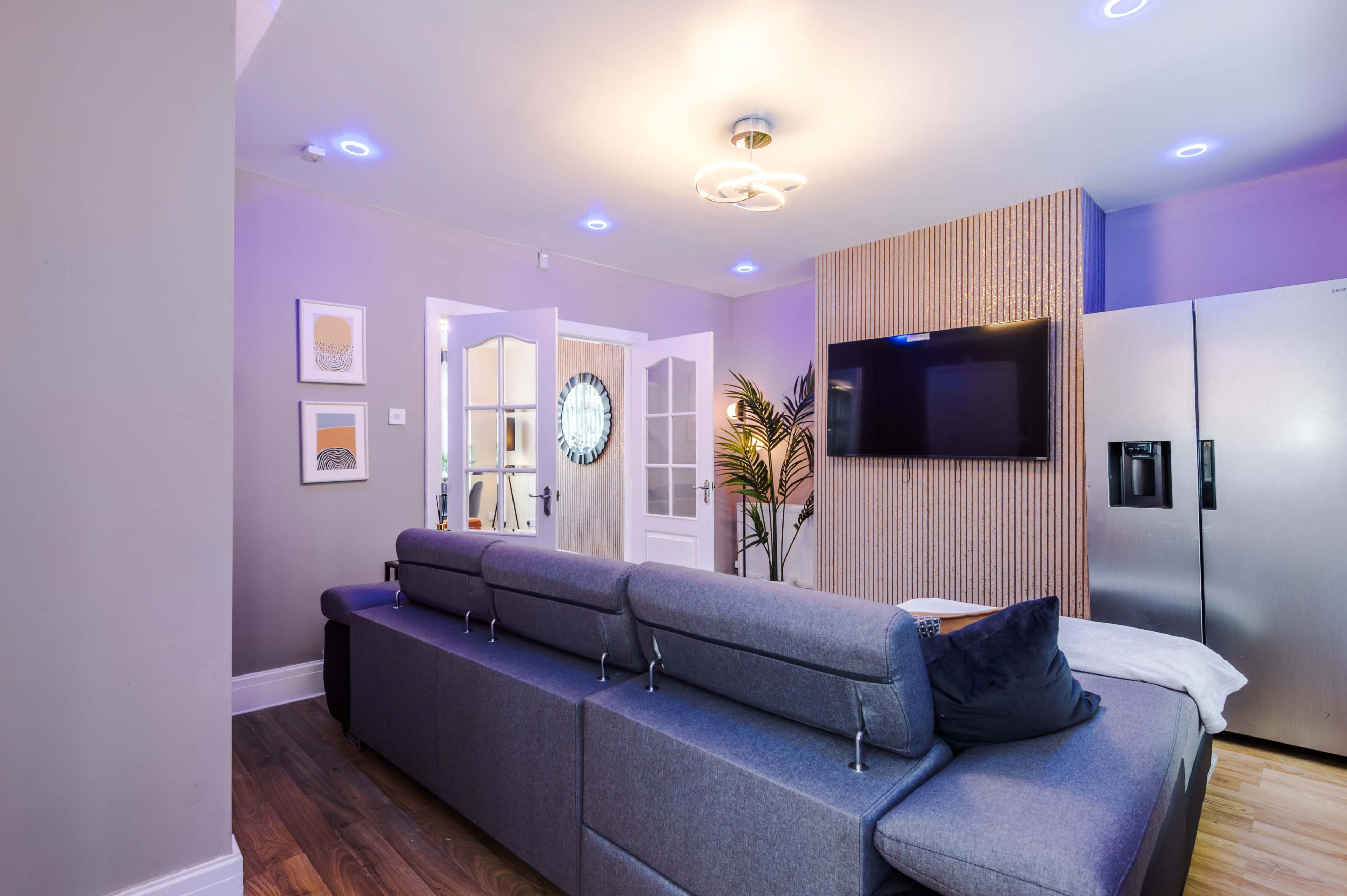 The image shows a modern living room with a gray sectional sofa, a mounted television, a refrigerator, and a decorative plant near the entrance.