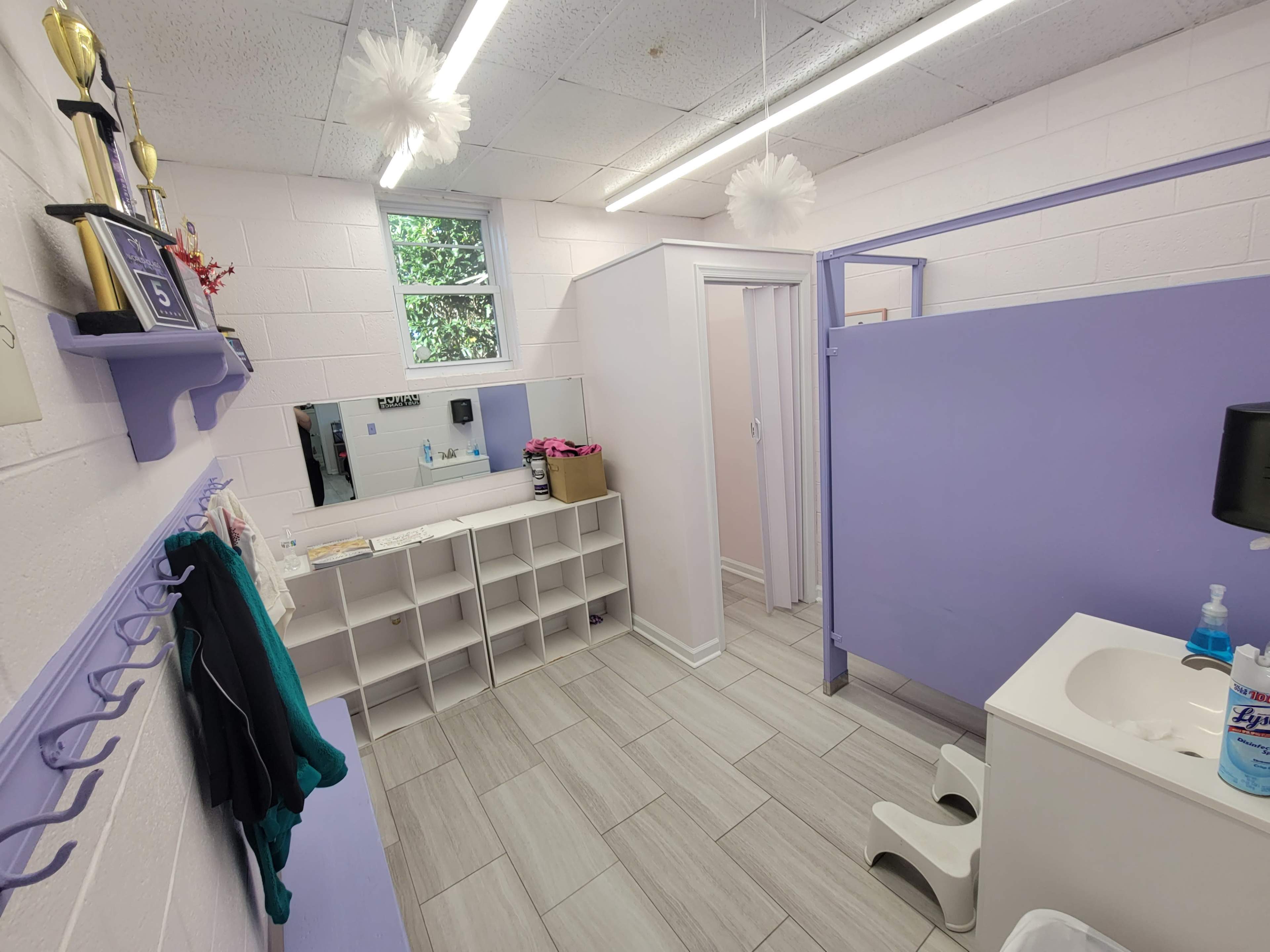 A lavender-colored bathroom with shelving, a mirror, and a shower stall, featuring tile flooring and a small sink.