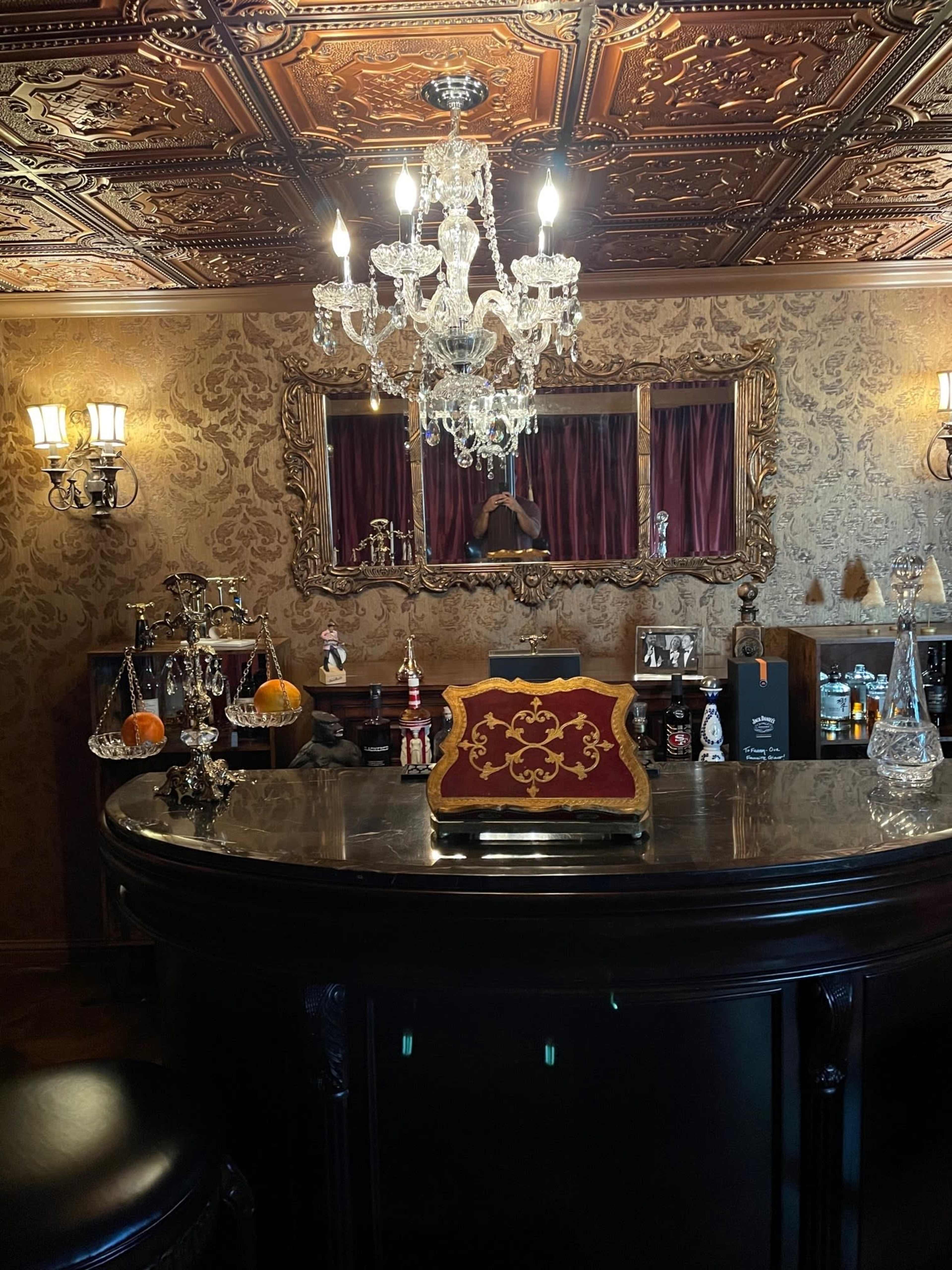 A well-appointed home bar with a chandelier, ornate wall decor, and an elegantly styled counter displaying various bottles and glassware.