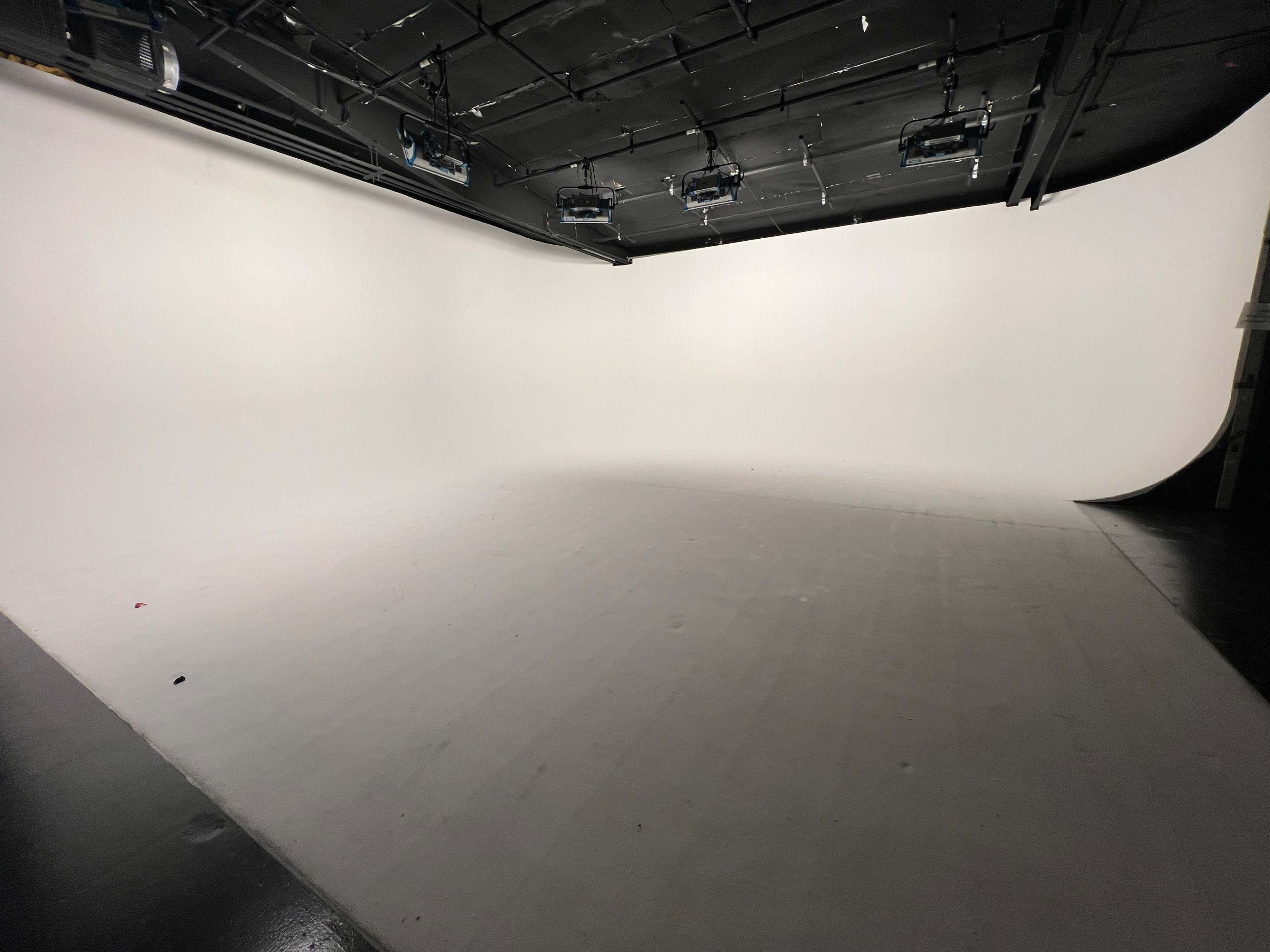 The image shows a large, empty photography studio with a seamless white backdrop and overhead lighting.
