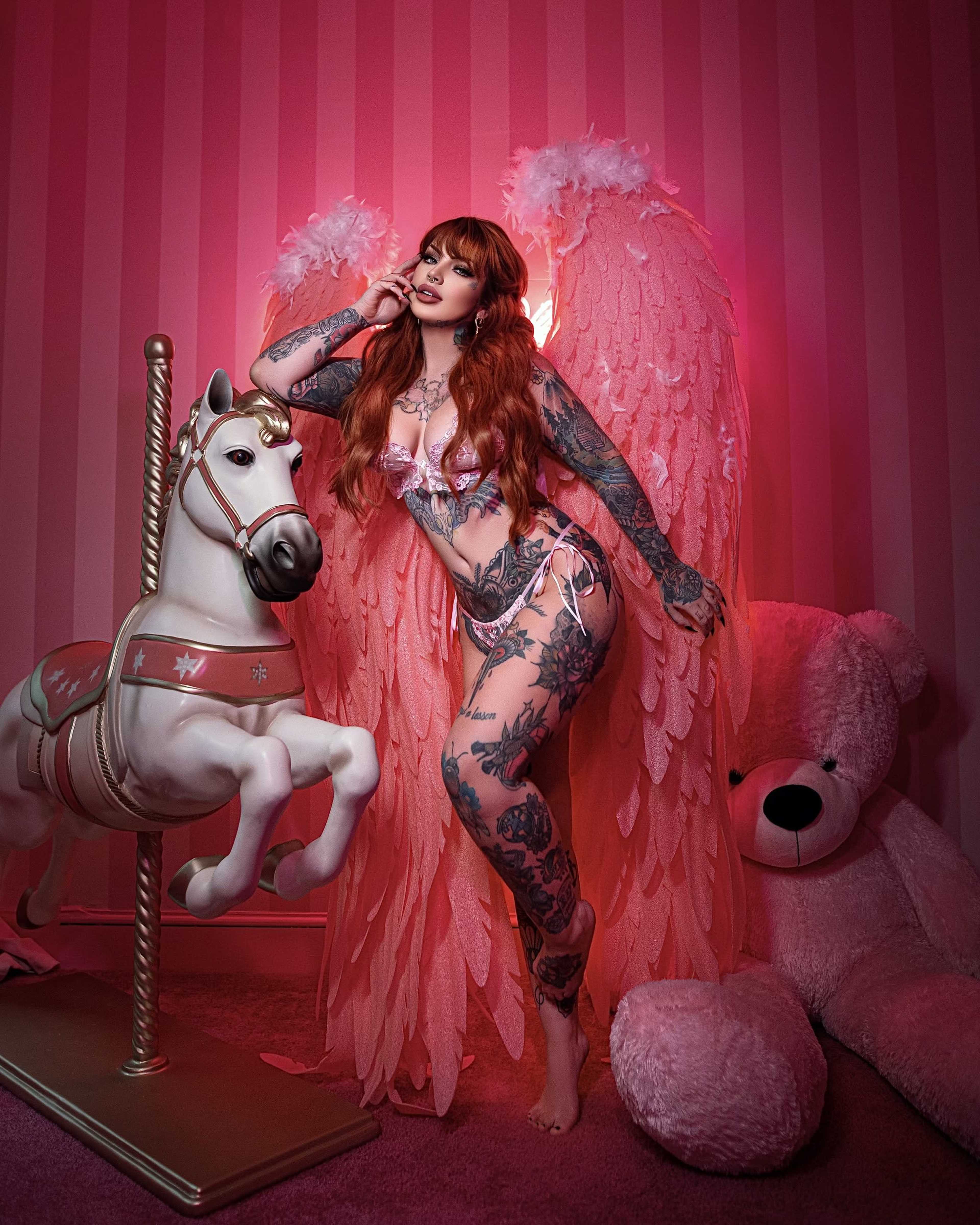 A woman with large feathered wings posed next to a white carousel horse and a large stuffed bear in a pink-striped room.