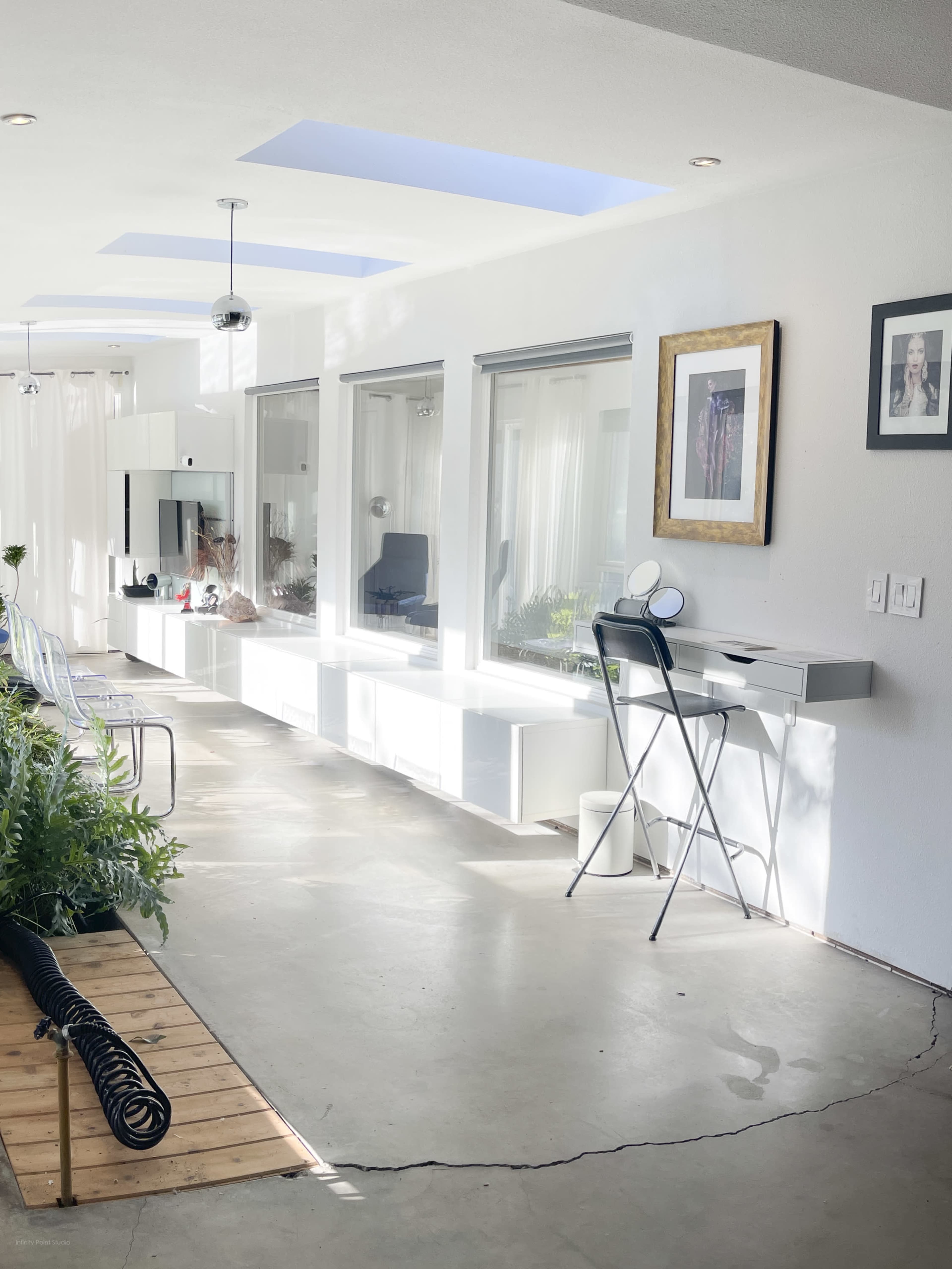 The image depicts a bright, contemporary room with a long, white wall cabinet and a small workspace featuring a chair and table, surrounded by plants and natural light.