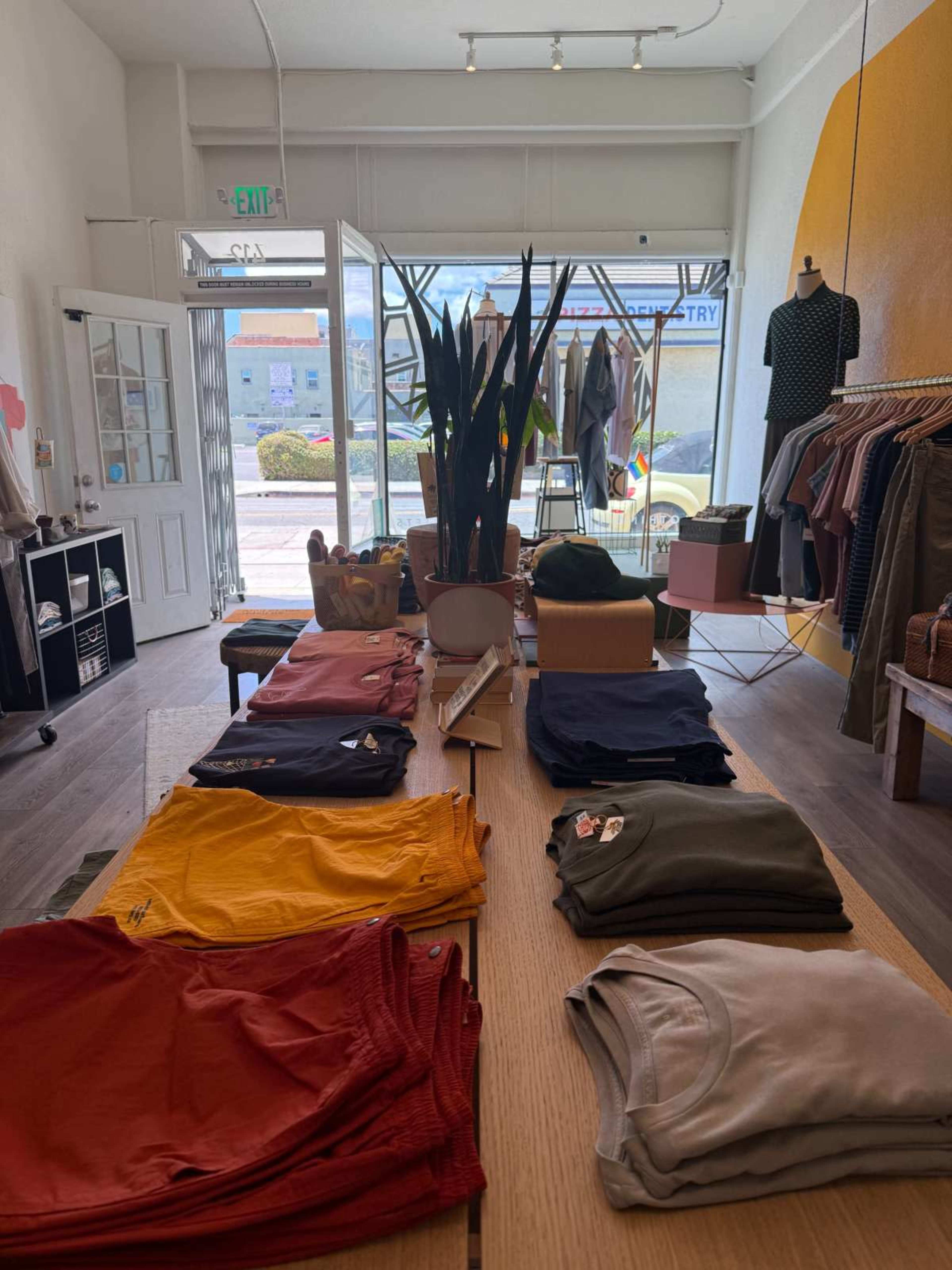 The image shows a clothing store interior with a display table featuring neatly arranged stacks of colored shirts and pants, surrounded by clothing racks and a large window displaying the outside.