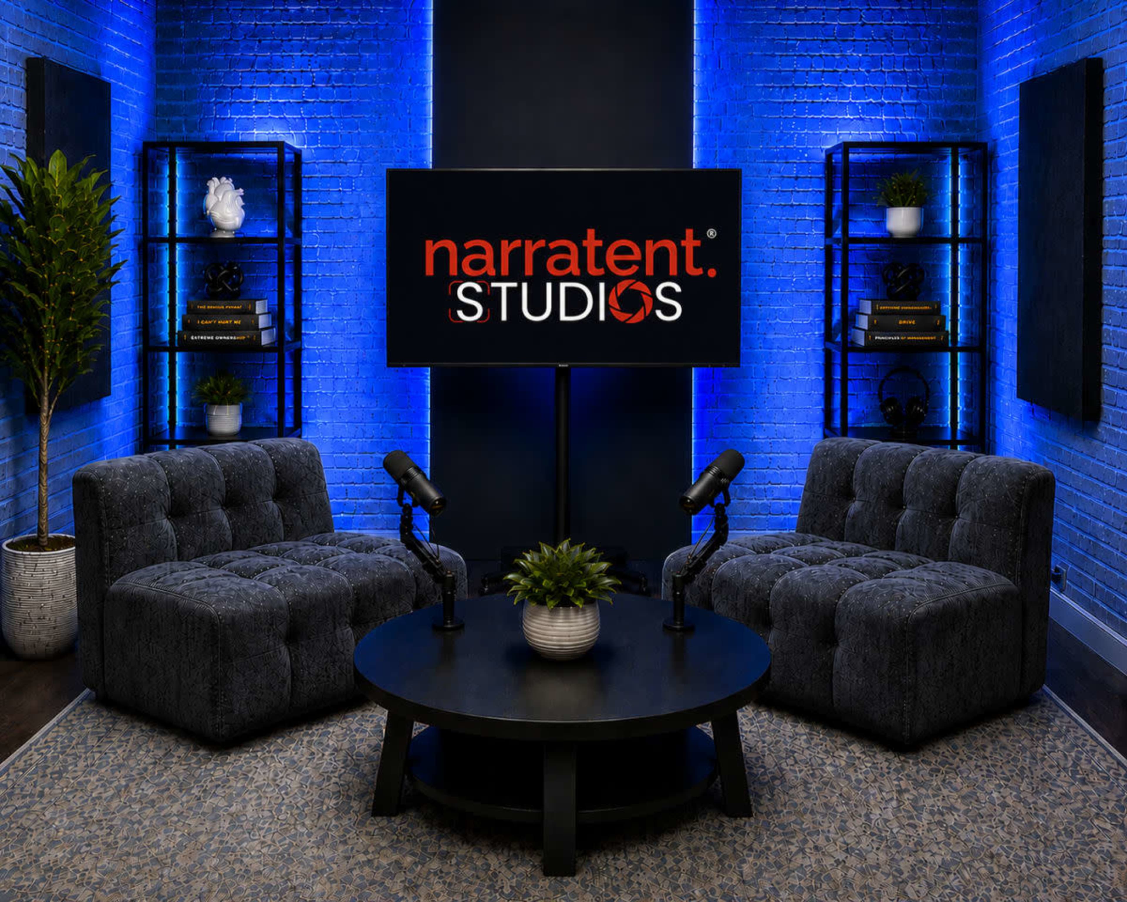 The image shows a modern podcast studio setup with two tufted chairs, a round table with a potted plant, and a large screen displaying "narratent STUDIOS," all set against a backdrop of blue-lit walls and shelves.