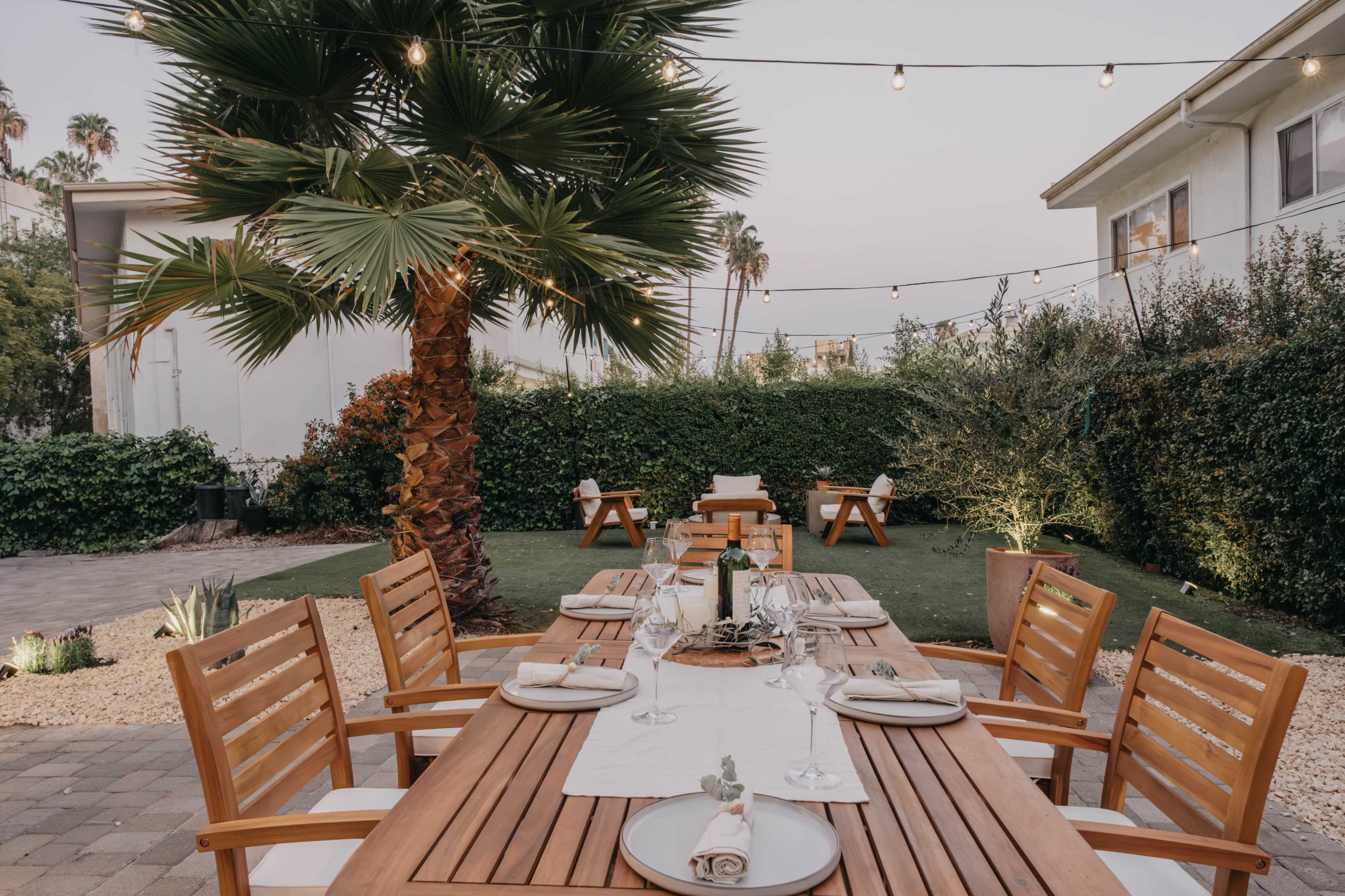 A wooden dining table is set for dinner in a well-lit outdoor garden featuring a palm tree and string lights overhead.