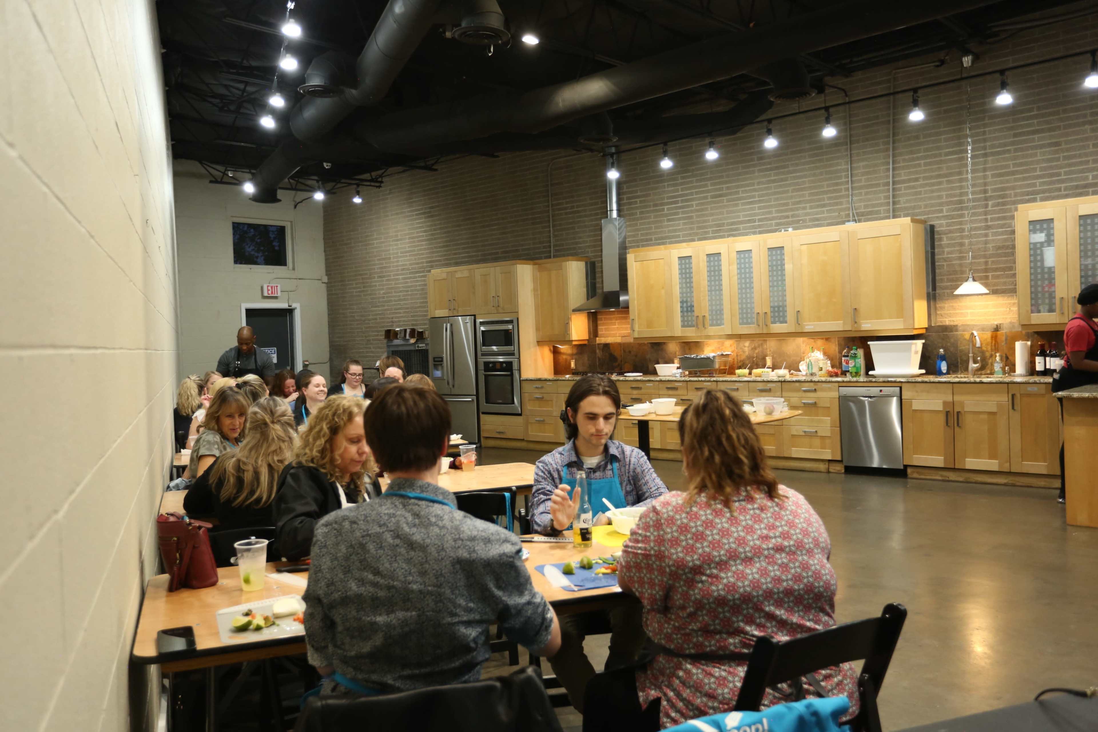 A group of people sits at tables in a kitchen space, engaged in food preparation or cooking activities, while a chef demonstrates at the front.
