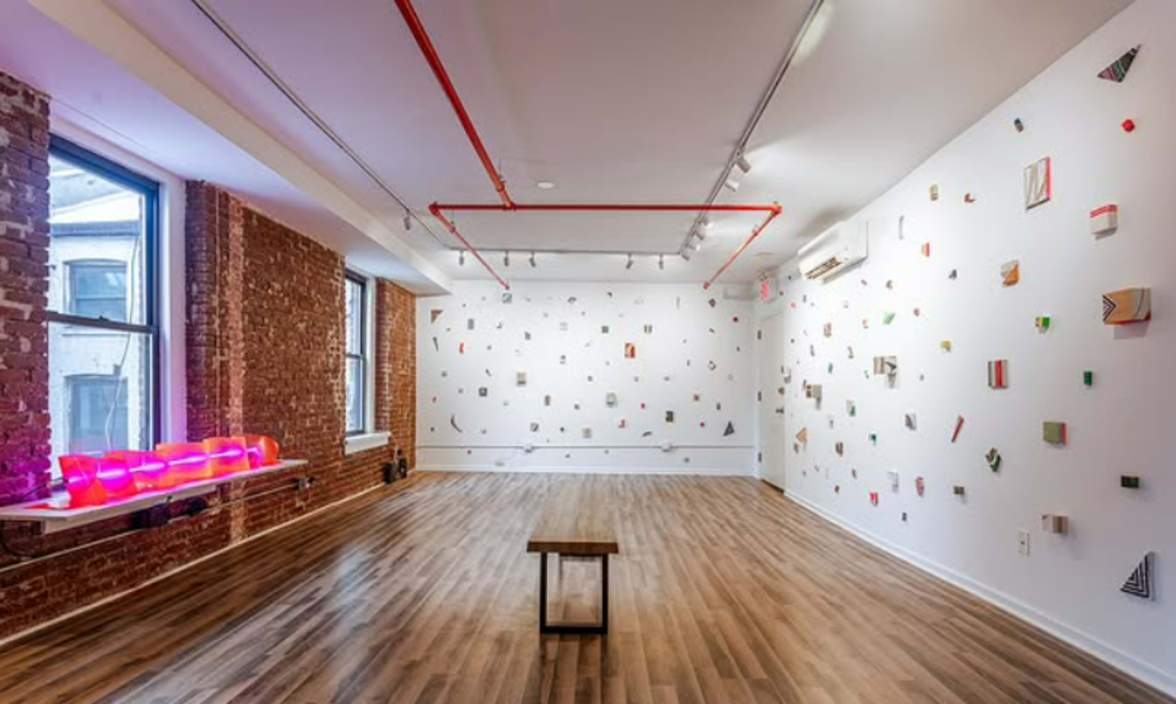 The image depicts a spacious art gallery with a wooden floor, a simple table in the center, and a white wall adorned with various colorful geometric art pieces.