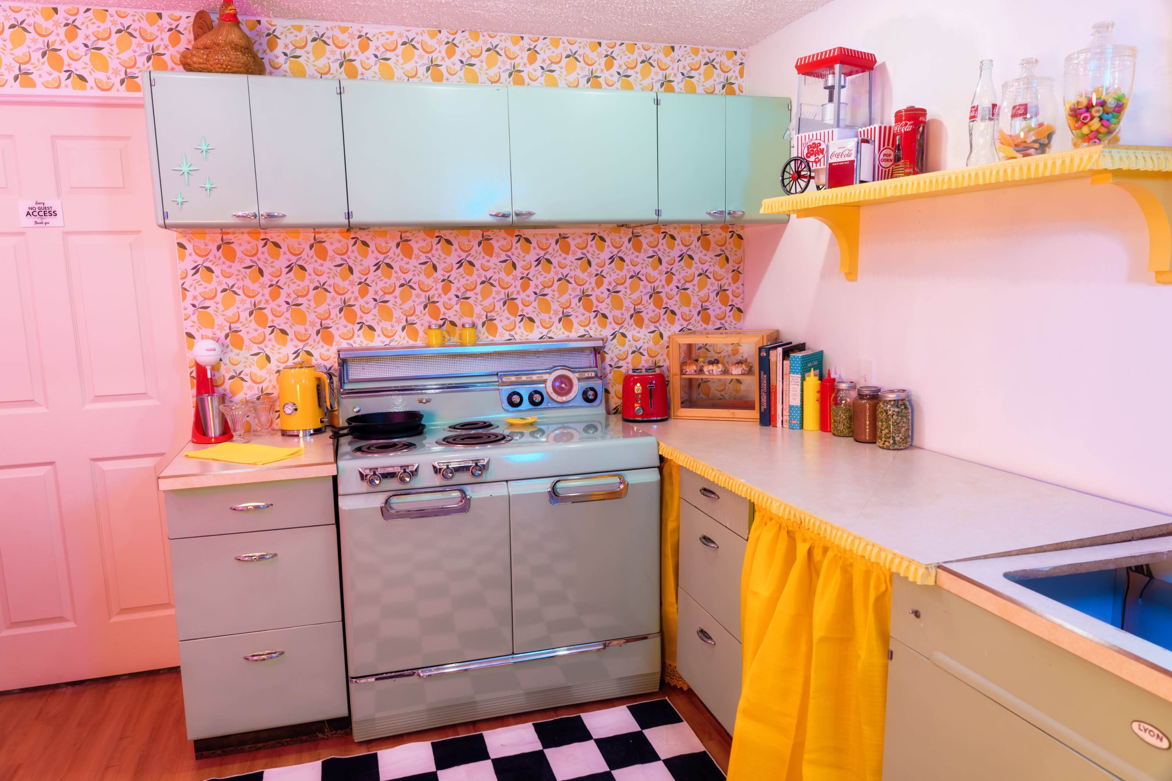 The image shows a vintage kitchen with mint green cabinets, a checkered floor, and retro appliances, all featuring a lemon-themed wallpaper.