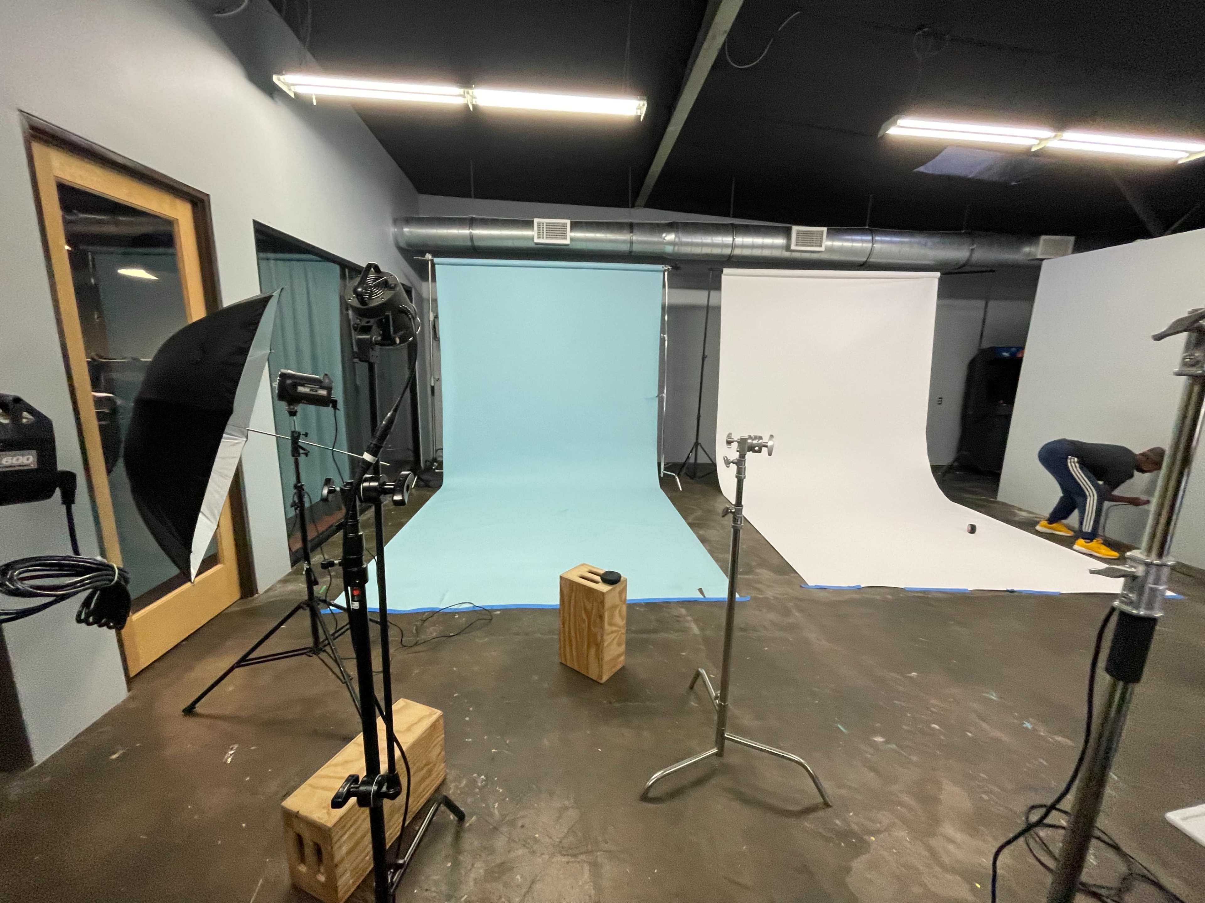A photography studio with a light blue backdrop on one side and a white backdrop on the other, along with lighting equipment and a person in the background.