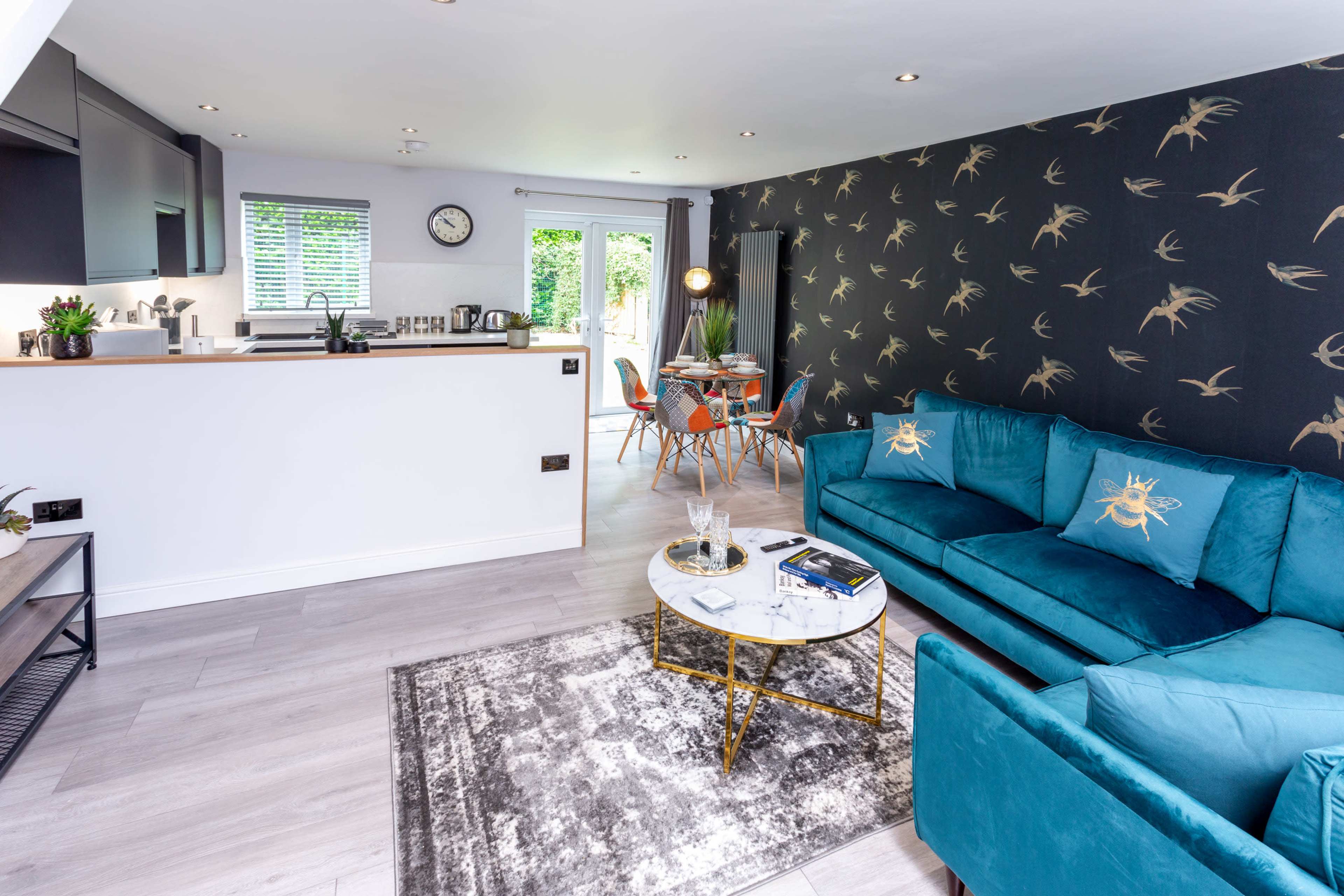 A modern living area features a teal sofa, a round coffee table, and a dining space with colorful chairs, all set against a patterned wall.