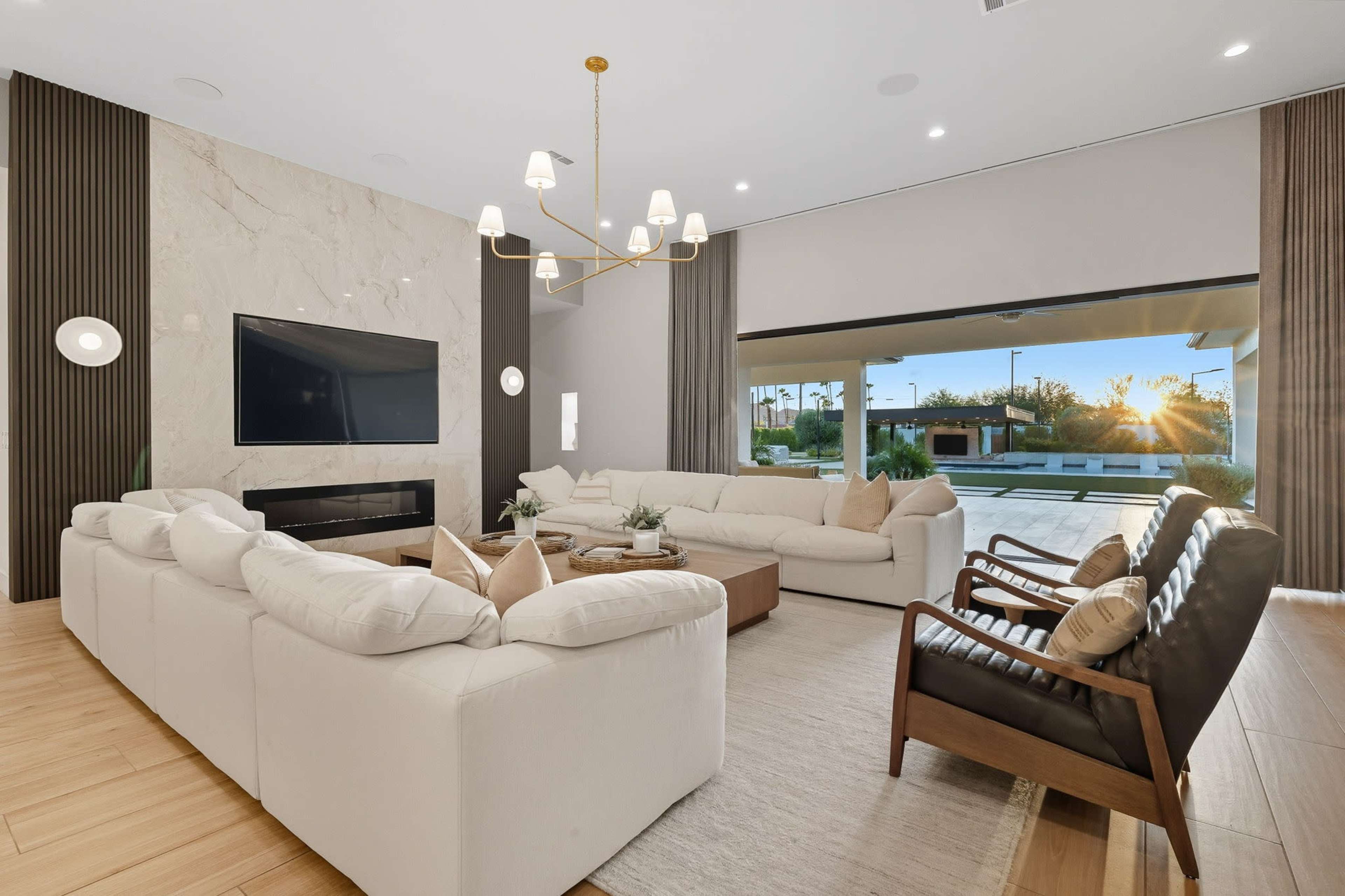 A spacious living room features a large sectional sofa, a wooden coffee table, and a backdrop of sliding glass doors that open to an outdoor space with a sunset view.