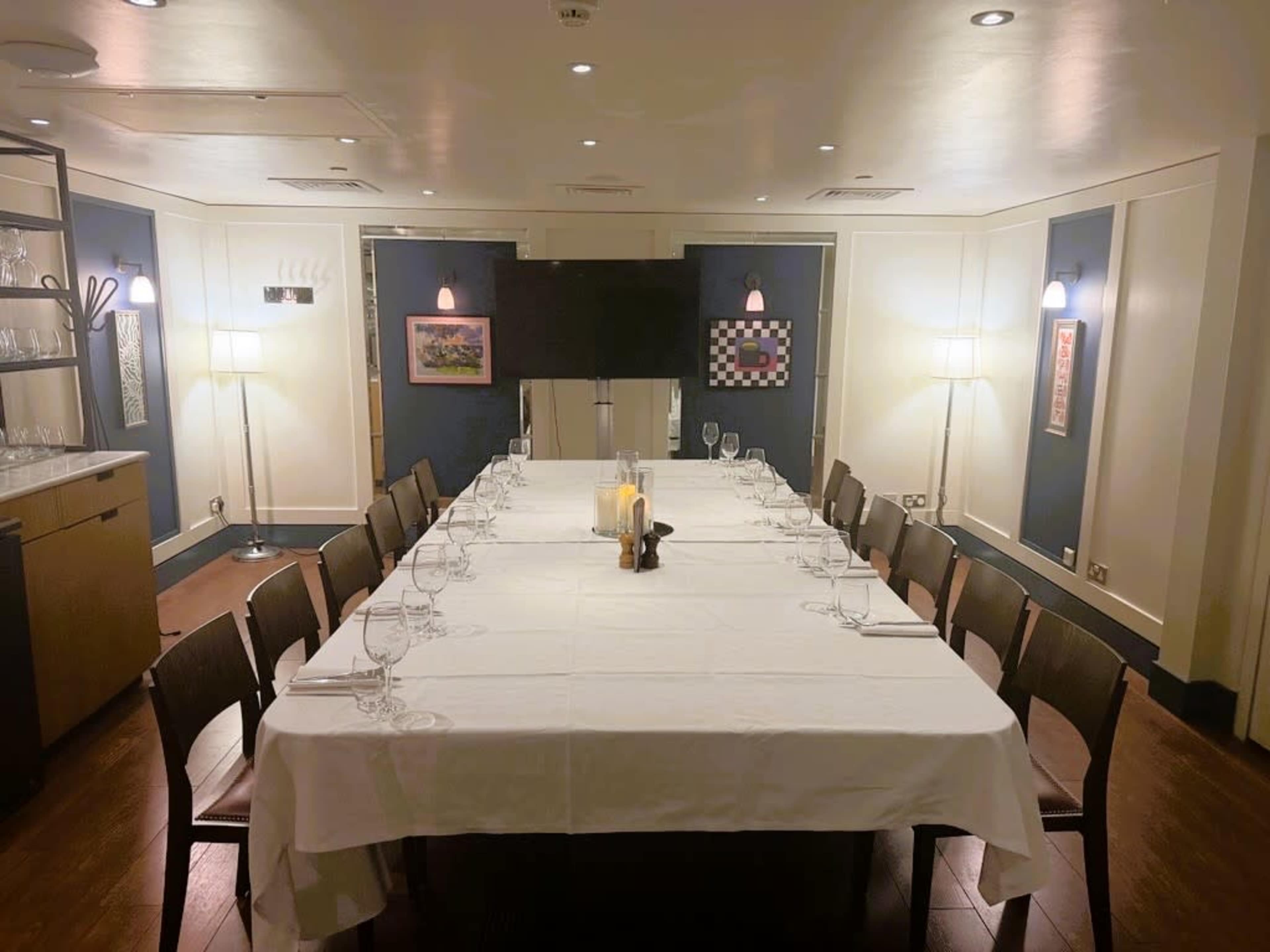 A long, rectangular dining table is set with glasses and a white tablecloth in a well-lit, private dining room.