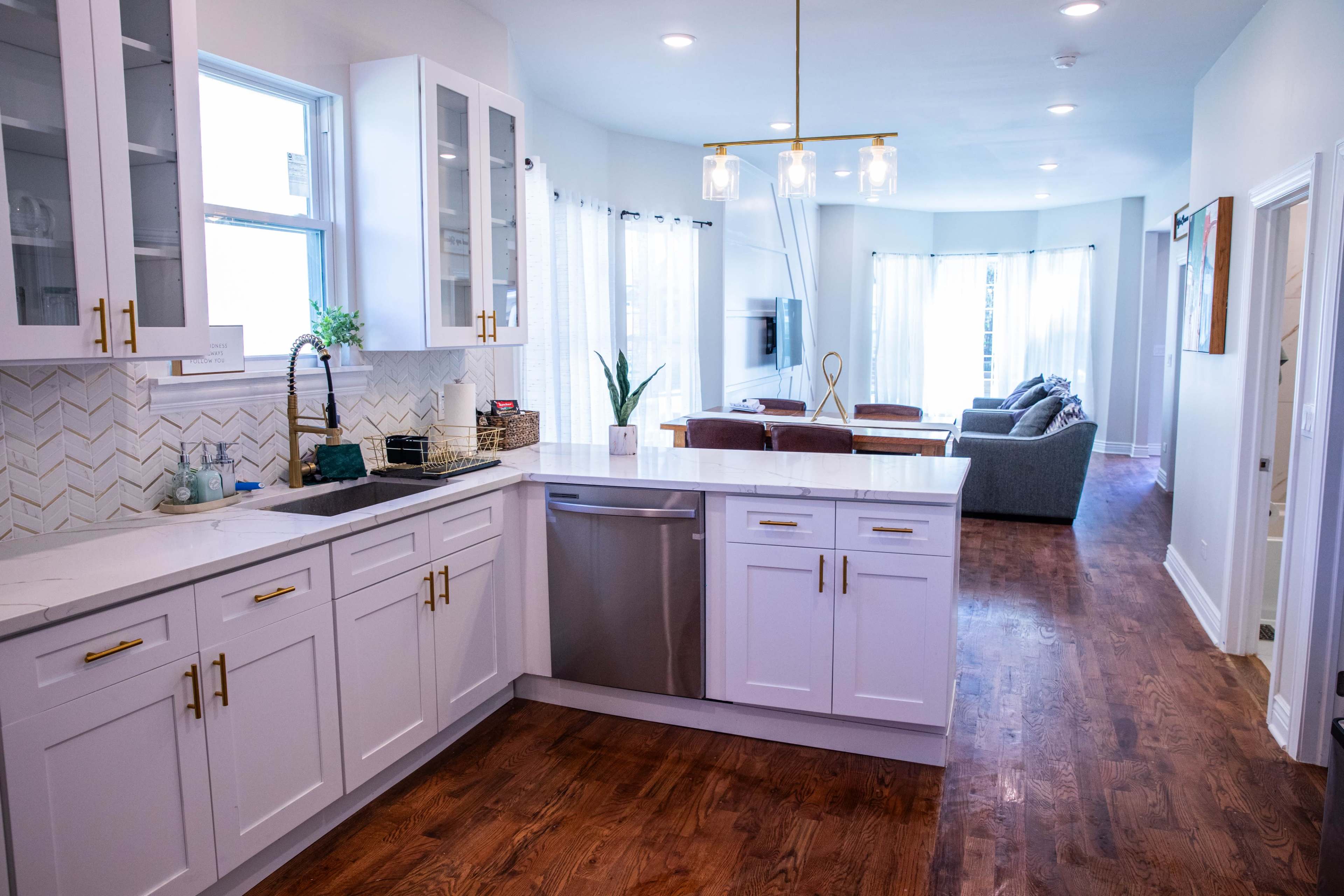 The image shows a modern kitchen with white cabinets and a marble countertop, adjacent to a living area with a sofa and large windows.