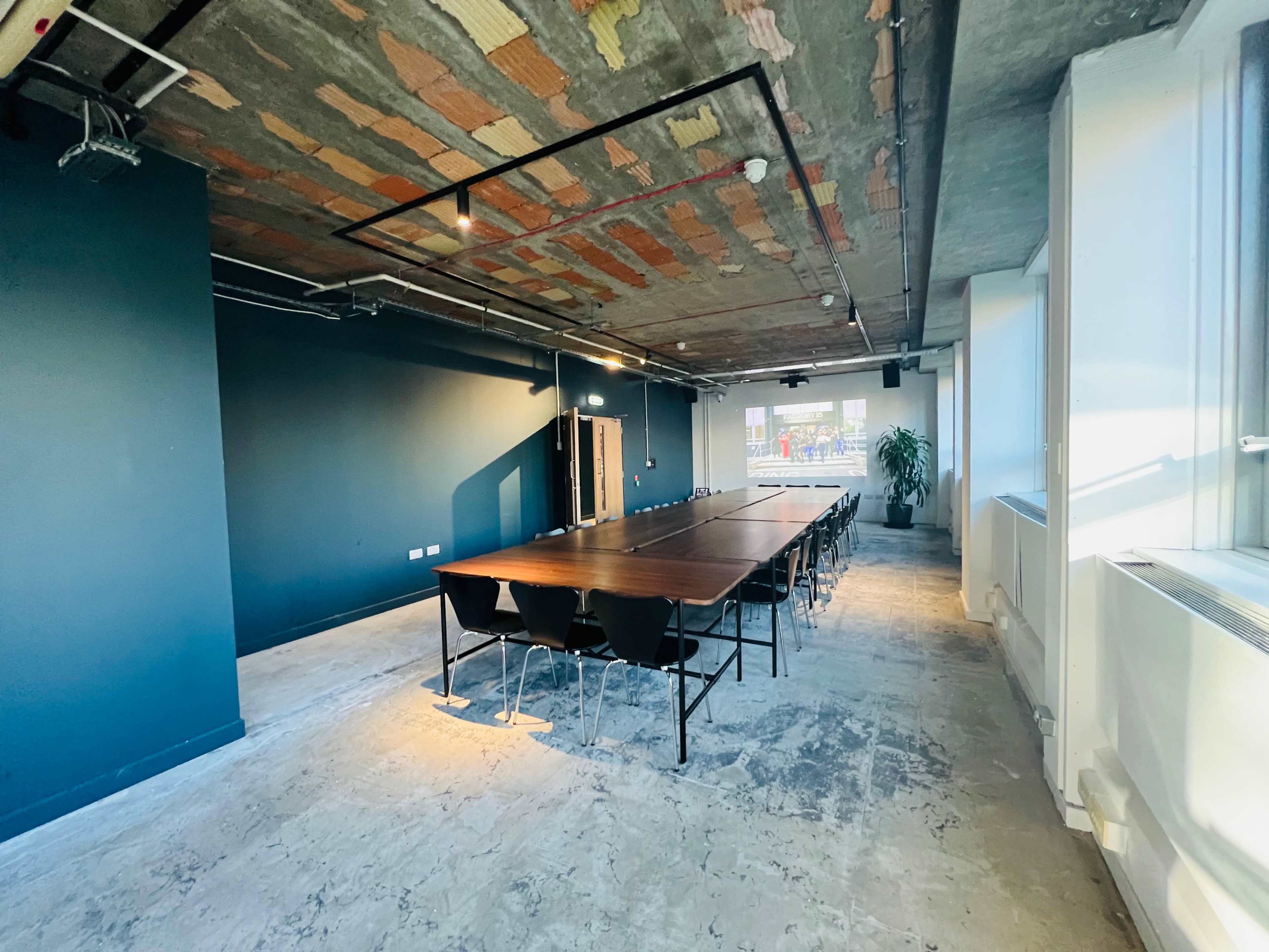 The image shows a large meeting room with a long wooden table and black chairs, featuring exposed brick and concrete walls, and large windows letting in natural light.