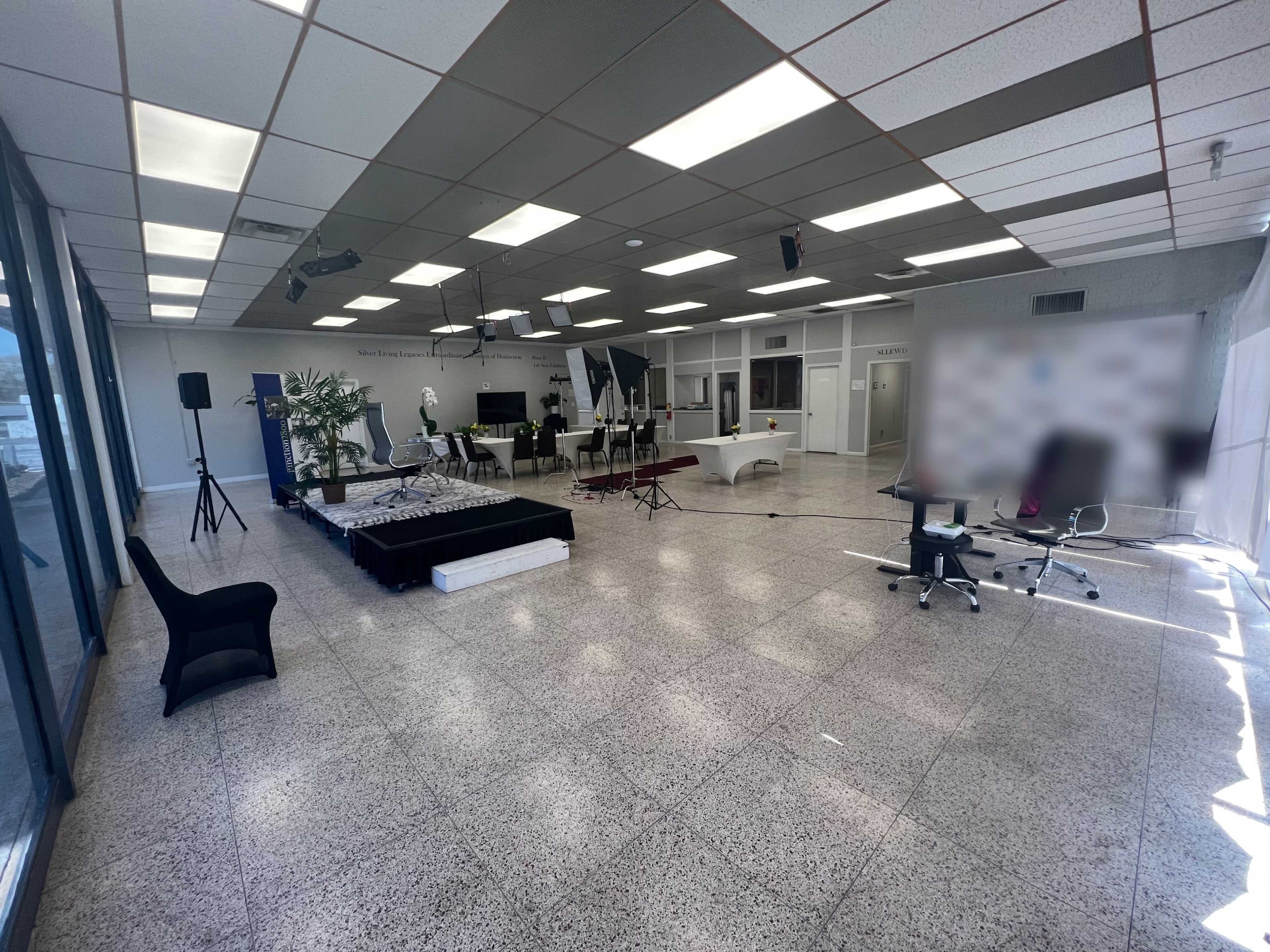 The image shows a spacious, well-lit interior of an office or event space set up with a seating area, a reception counter, and various audio-visual equipment.