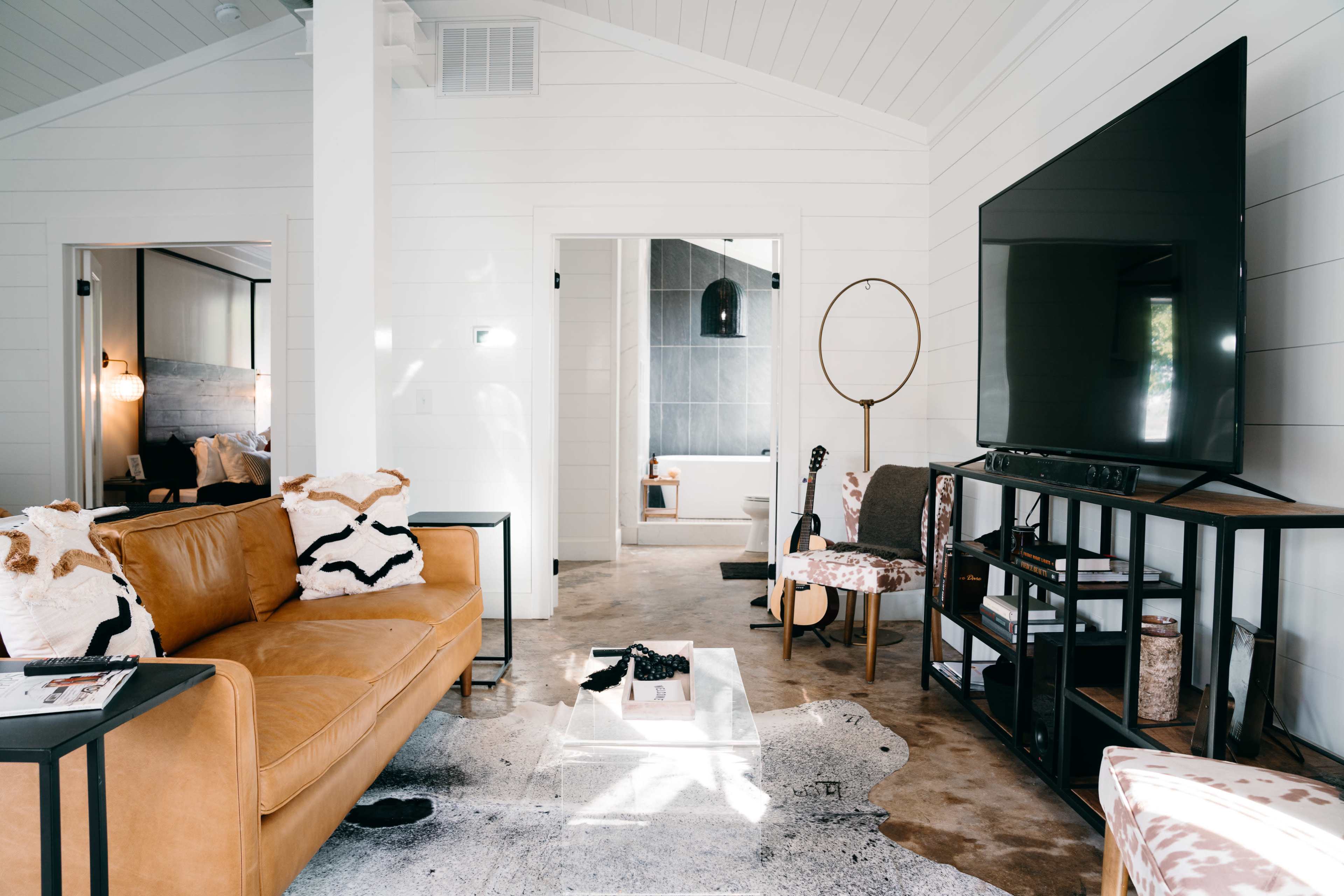 A cozy living room features a brown leather sofa, a large television on a black stand, and a decorative coffee table on a patterned rug.