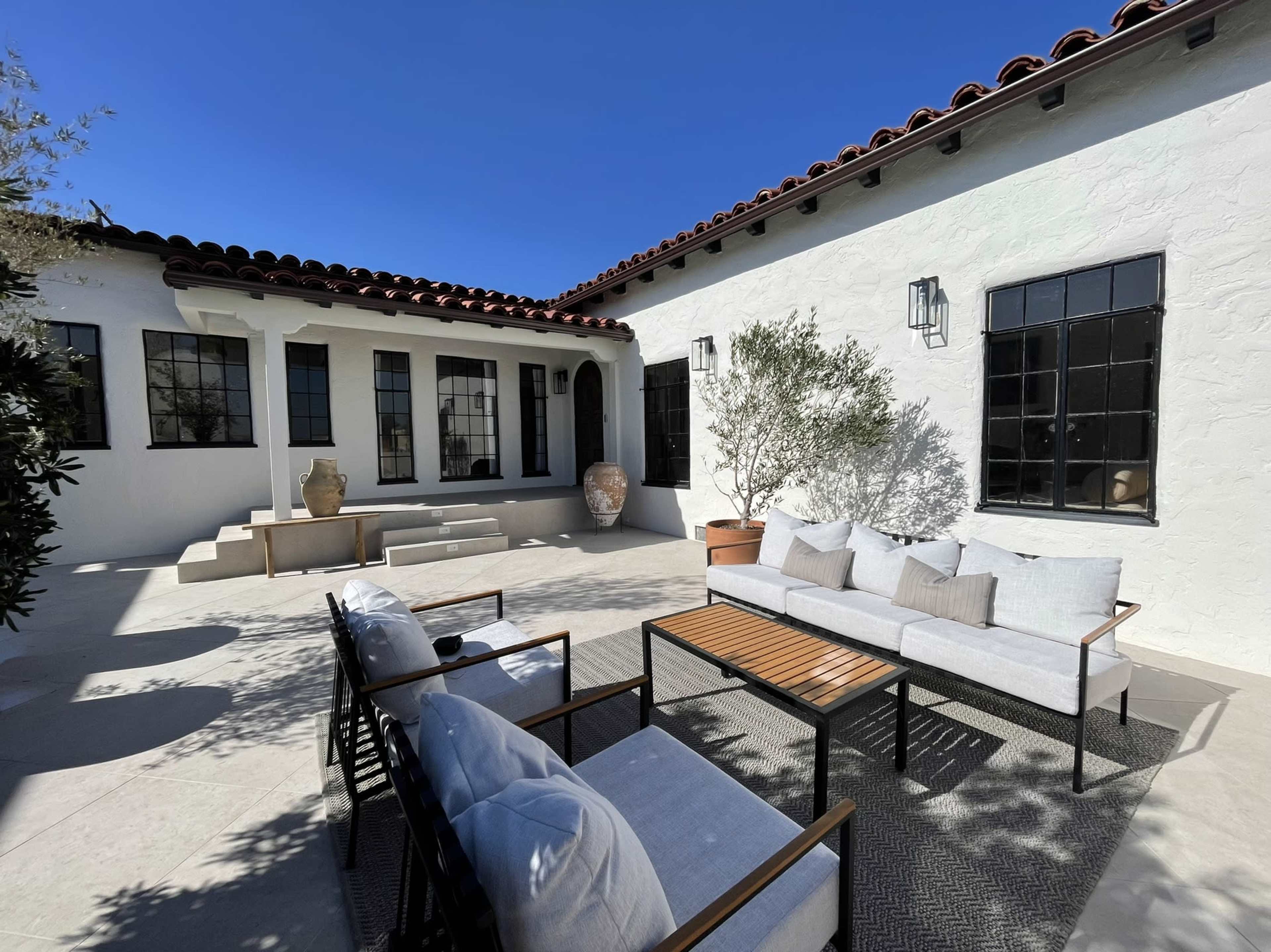 A patio with modern seating and a table is surrounded by white walls and large windows in a sunny courtyard.