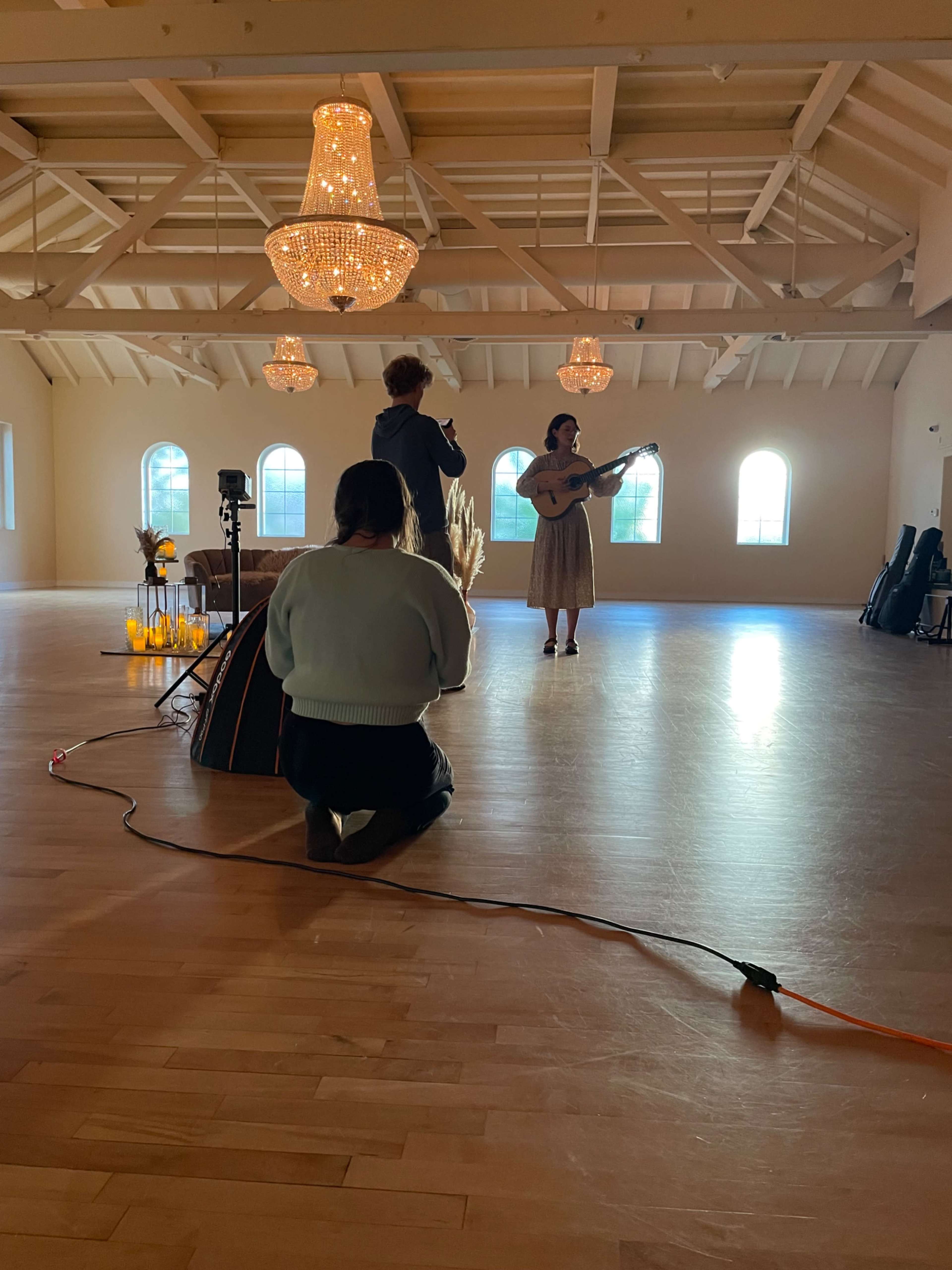 A woman plays a guitar in a spacious, well-lit room while another person records her performance, with decorative chandeliers overhead.