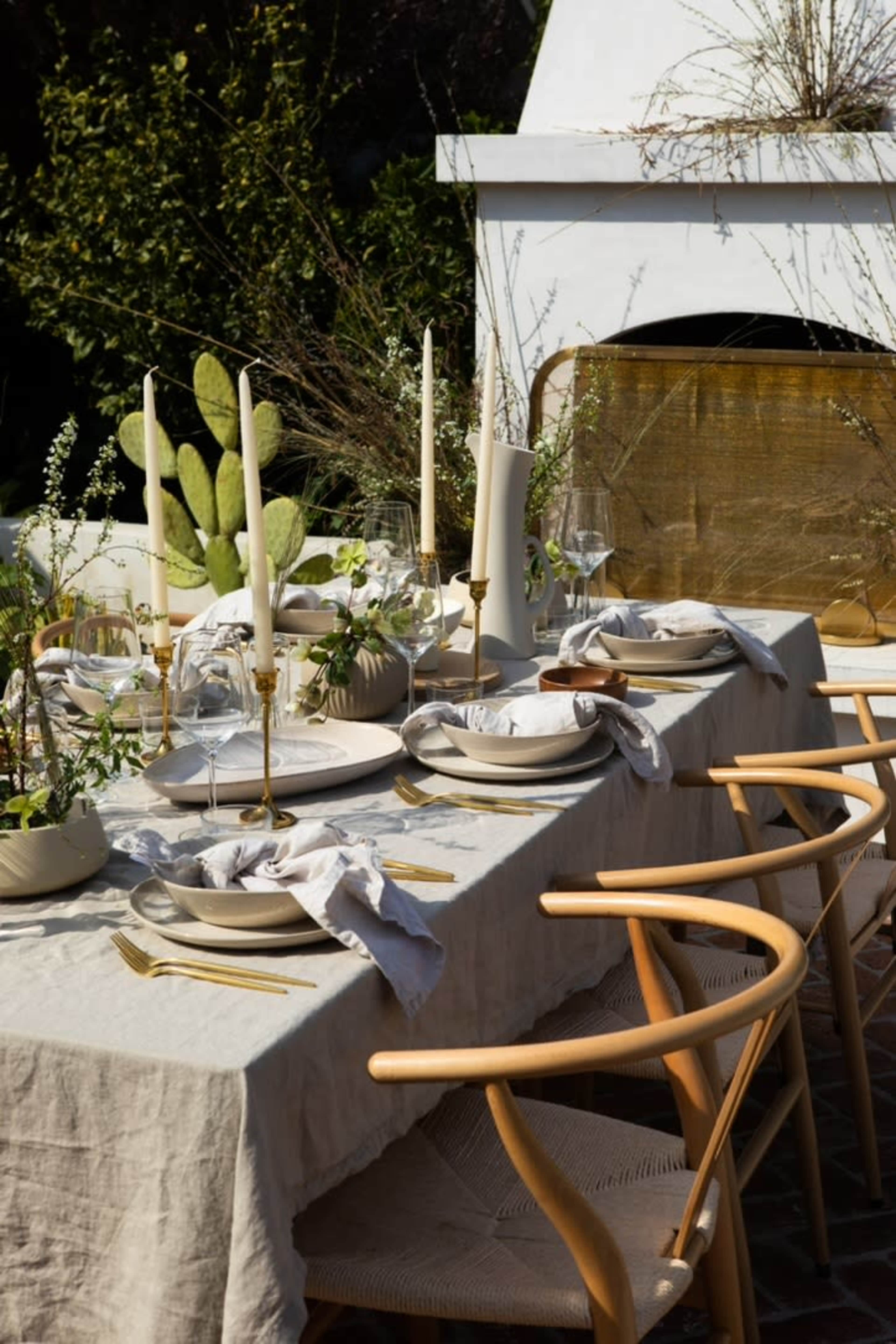 A beautifully arranged outdoor dining table is set with elegant dishes, glassware, and candlesticks, surrounded by greenery and a decorative backdrop.