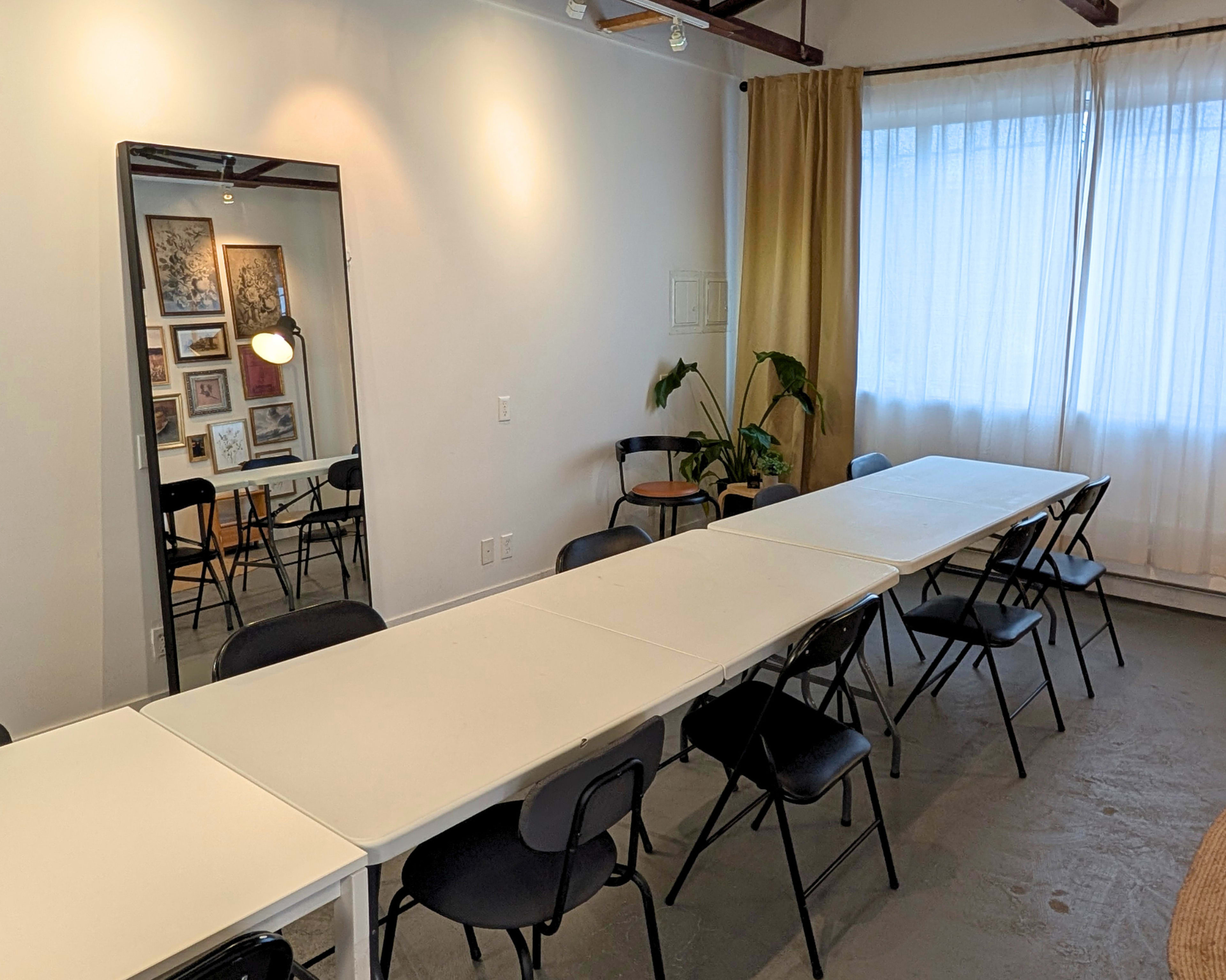 The image shows a meeting room with a long table surrounded by black chairs, a mirror on the wall, and large windows covered with sheer curtains.