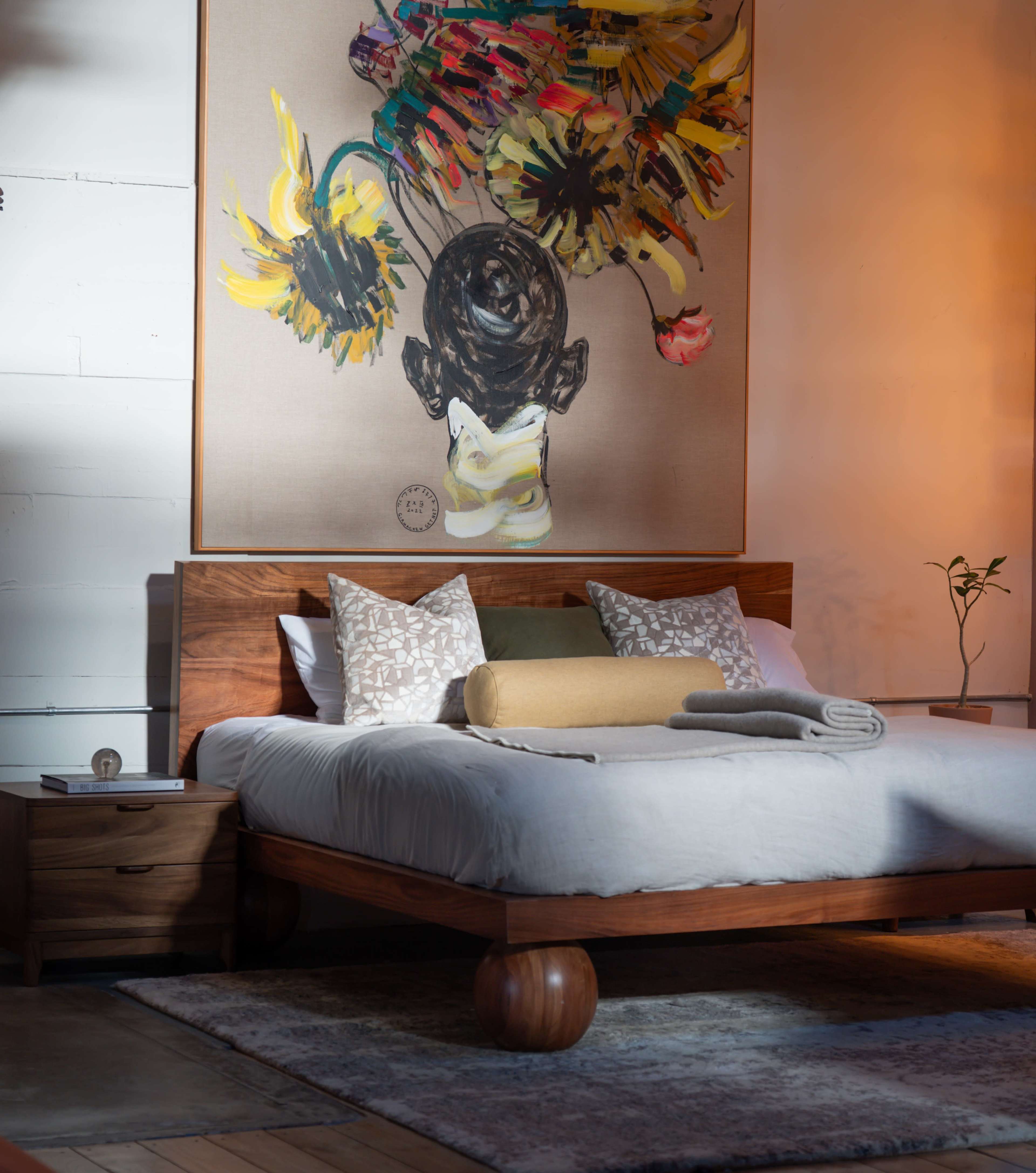 The image shows a modern bedroom featuring a wooden bed frame with decorative pillows, a large colorful artwork on the wall, and a small plant next to a nightstand.
