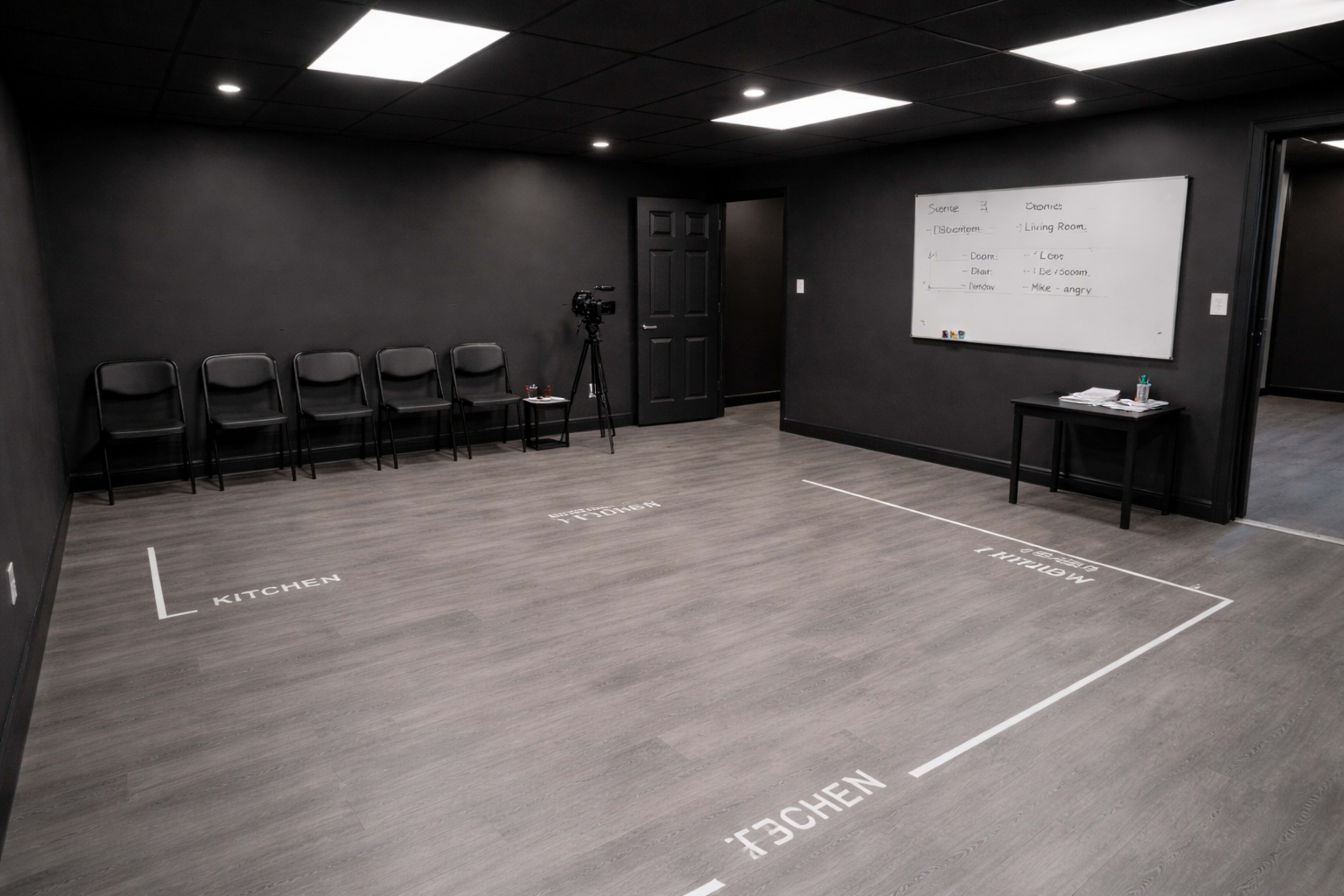 The image shows a dark room with a black wall, a set of empty chairs lined up against one wall, and a small table with supplies, labeled areas for "KITCHEN," "LIVING ROOM," and "DINING ROOM" on the floor.