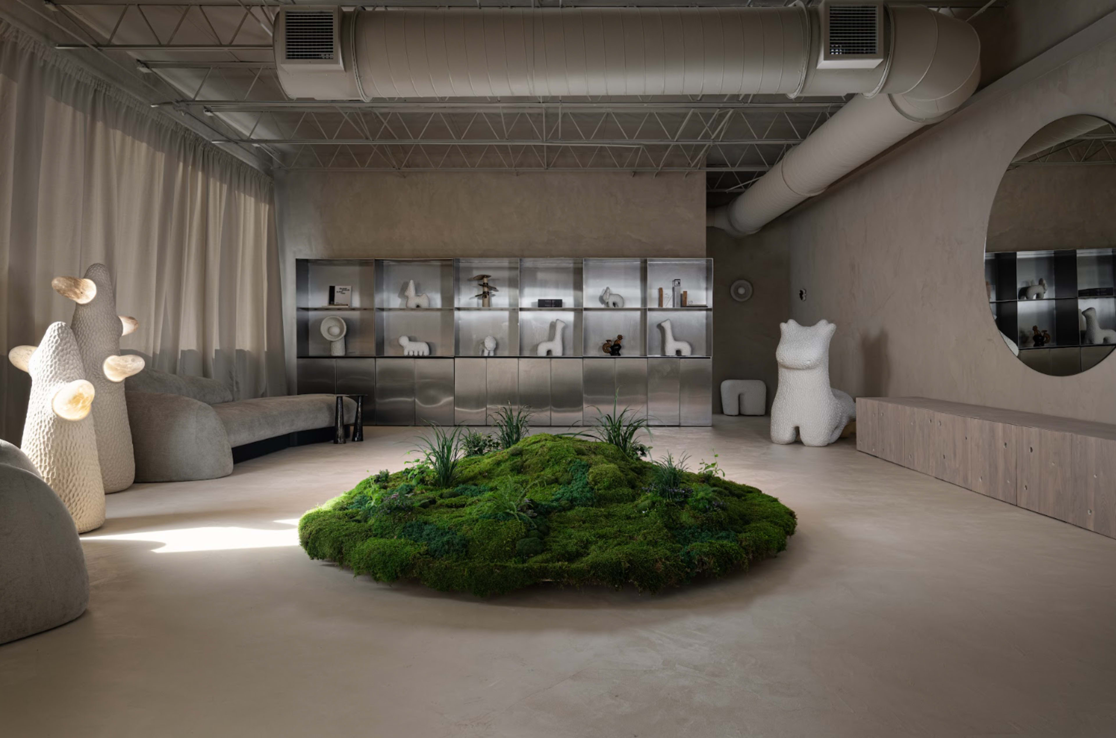 The image shows a modern interior space featuring a central moss-covered mound, surrounded by minimalist furniture and shelves displaying decorative items.