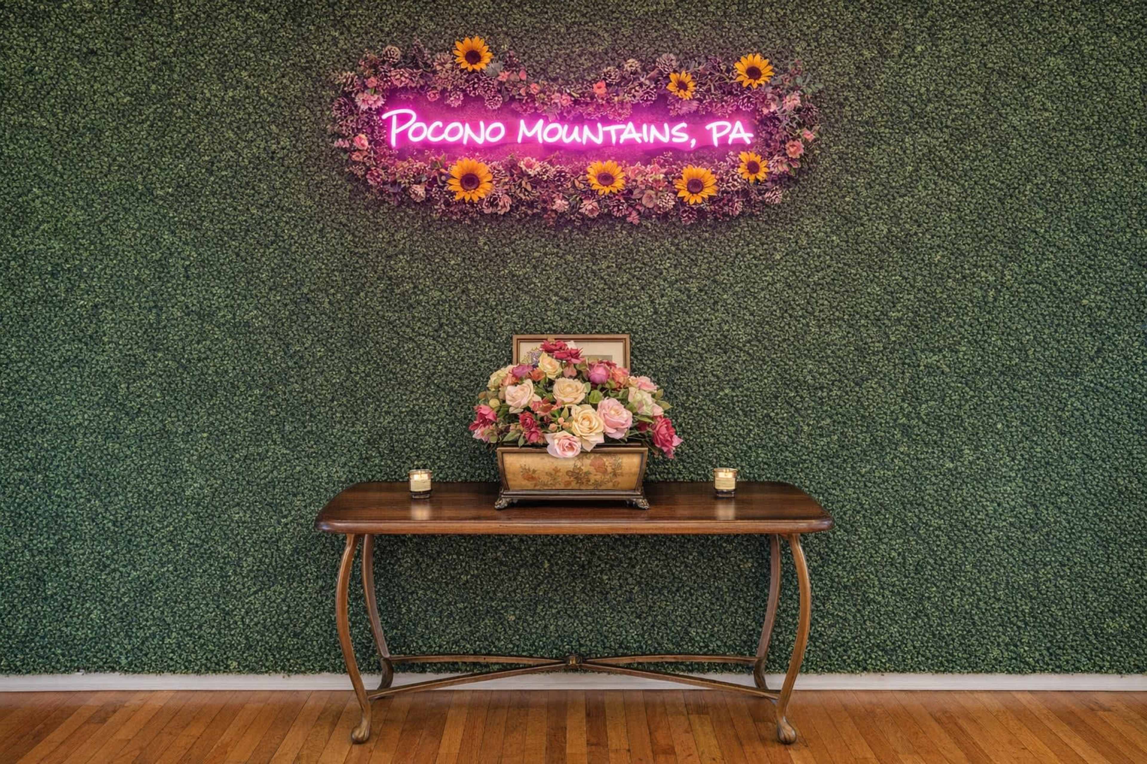 The image shows a wooden table adorned with a floral arrangement and candles, set against a green wall featuring a neon sign that reads "Pocono Mountains, PA."