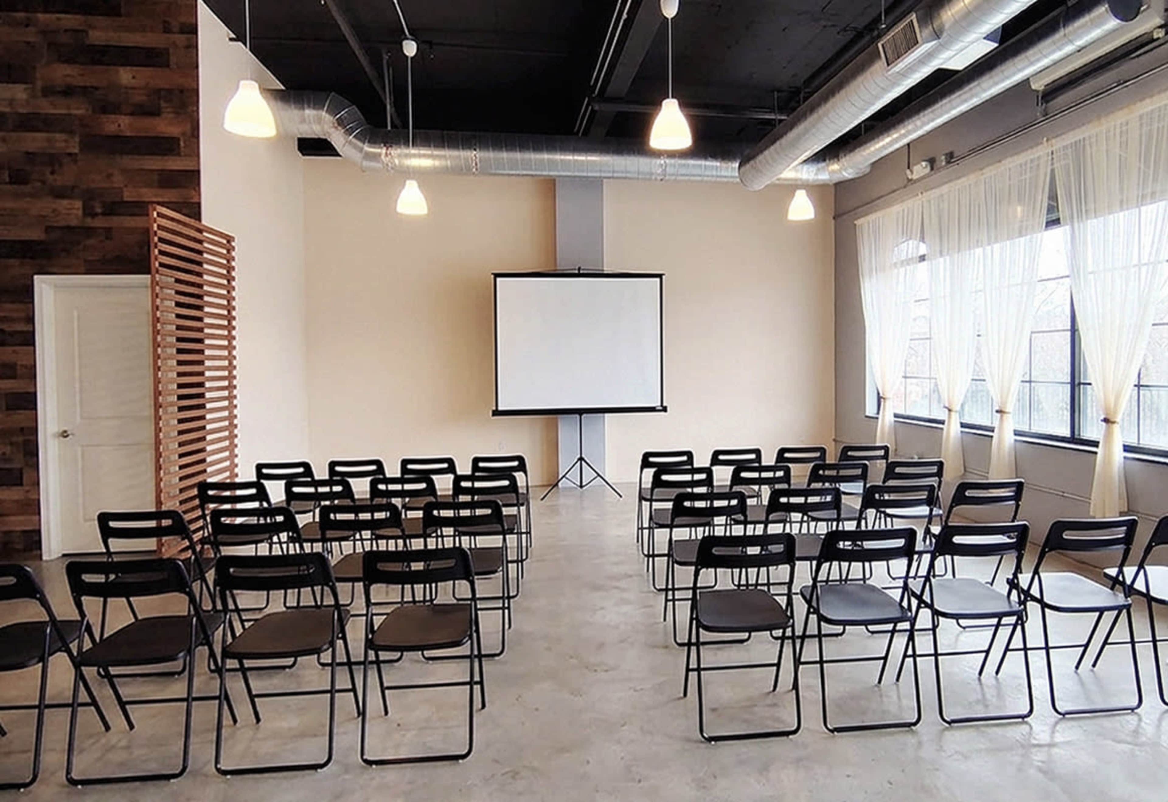 The image shows a spacious room arranged with rows of black folding chairs facing a blank projector screen, illuminated by overhead lights.