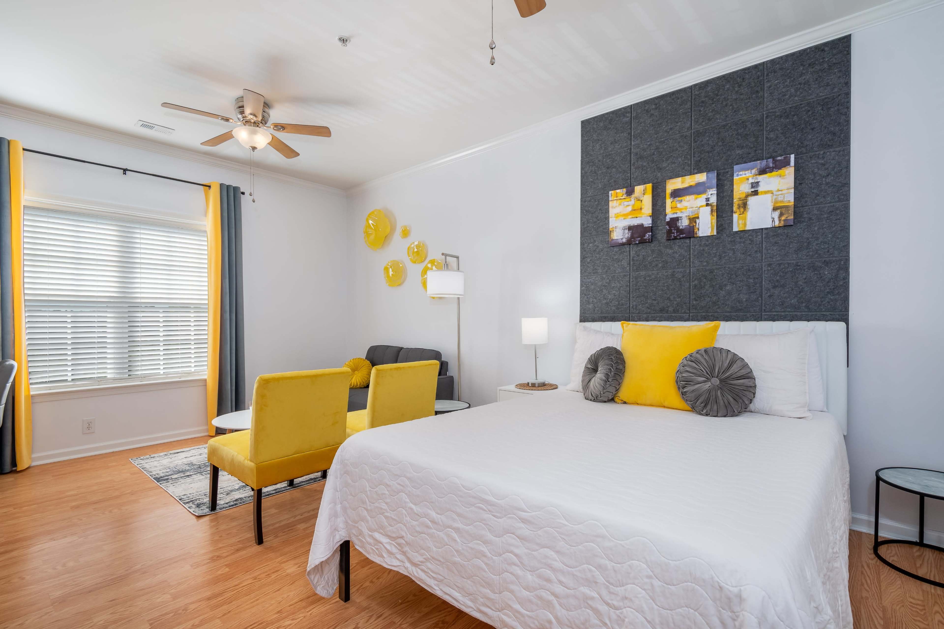 A bedroom with a bed, two yellow chairs, a small table, and decorative wall art.