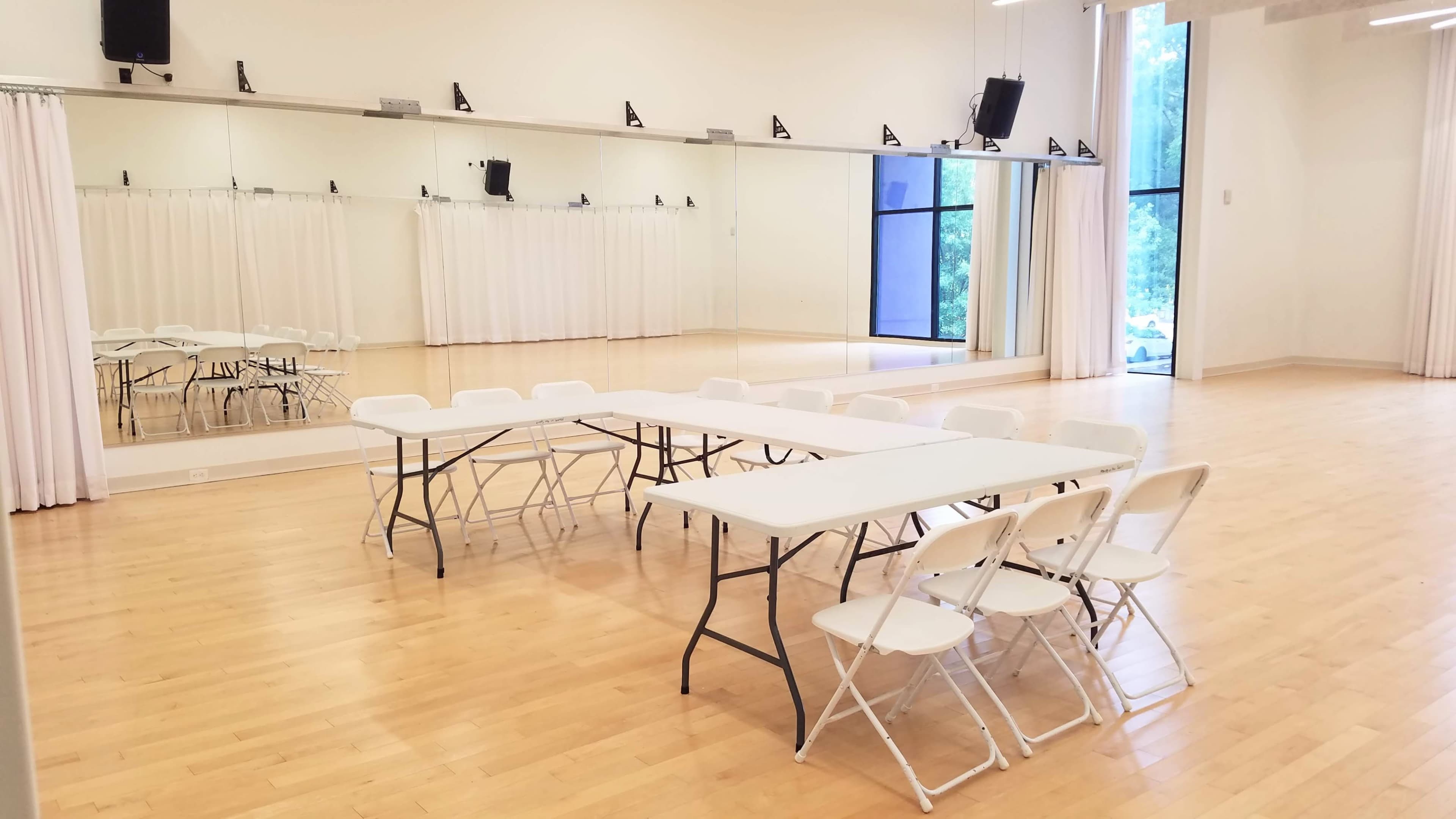 The image shows a spacious room with a hardwood floor, featuring a rectangular table surrounded by folding chairs and large mirrors along one wall.