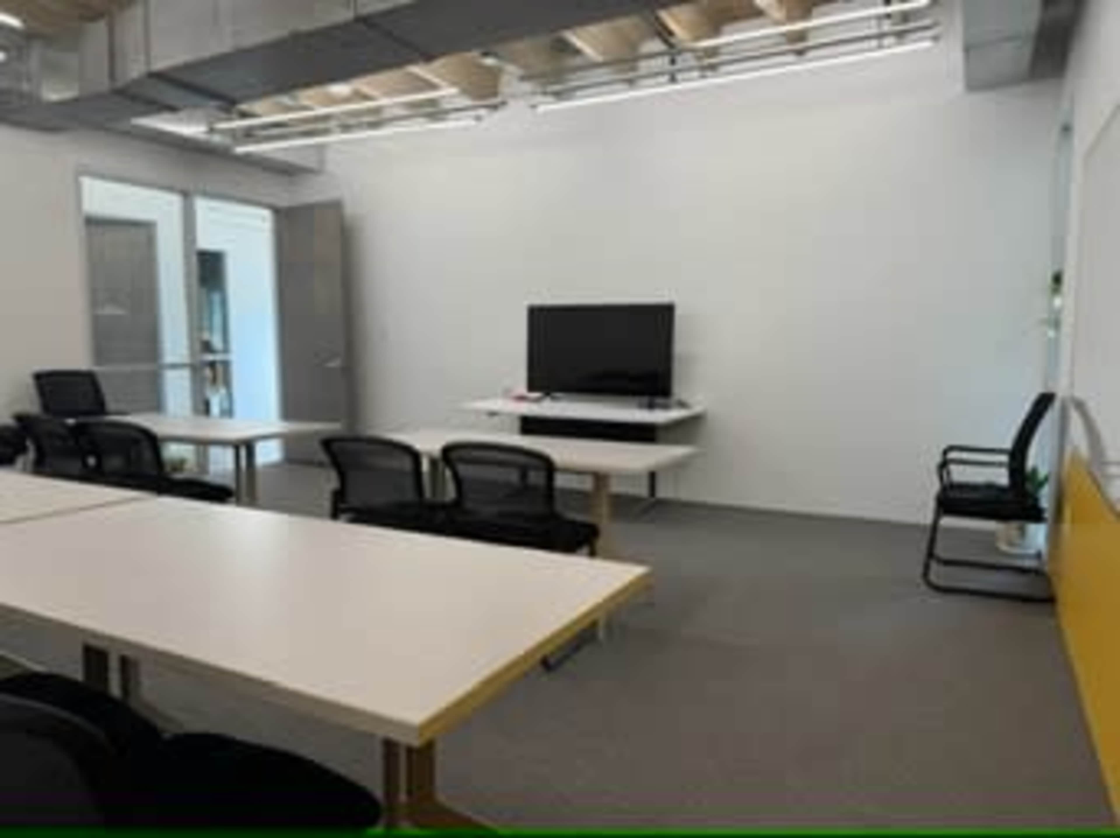 The image shows a modern office space with white walls, a large television mounted on a wall, multiple tables, and black chairs.