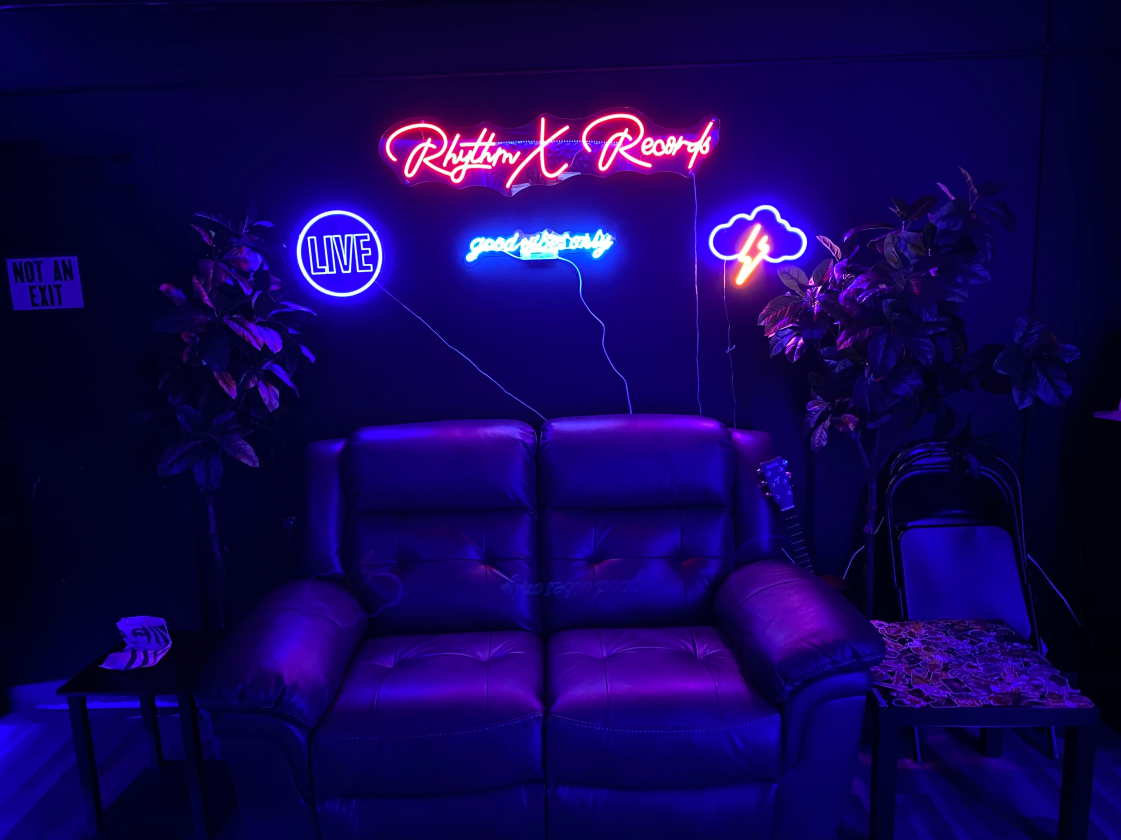 A leather couch against a dark wall adorned with colorful neon signs that read "Rhythm & Records," "LIVE," and "goodvibes."