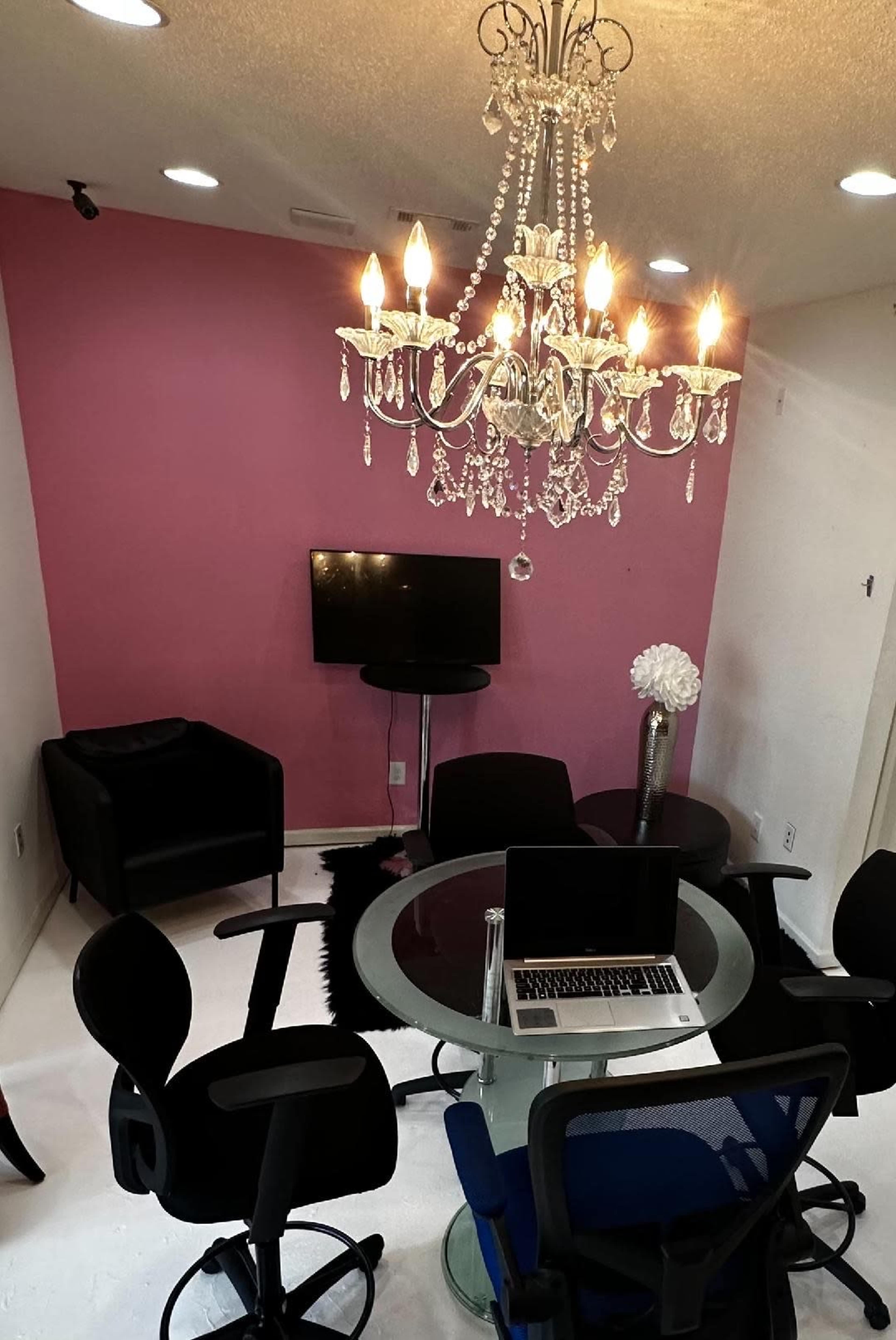 A meeting room features a round glass table surrounded by black and blue office chairs, illuminated by a chandelier, with a television mounted on a pink wall.